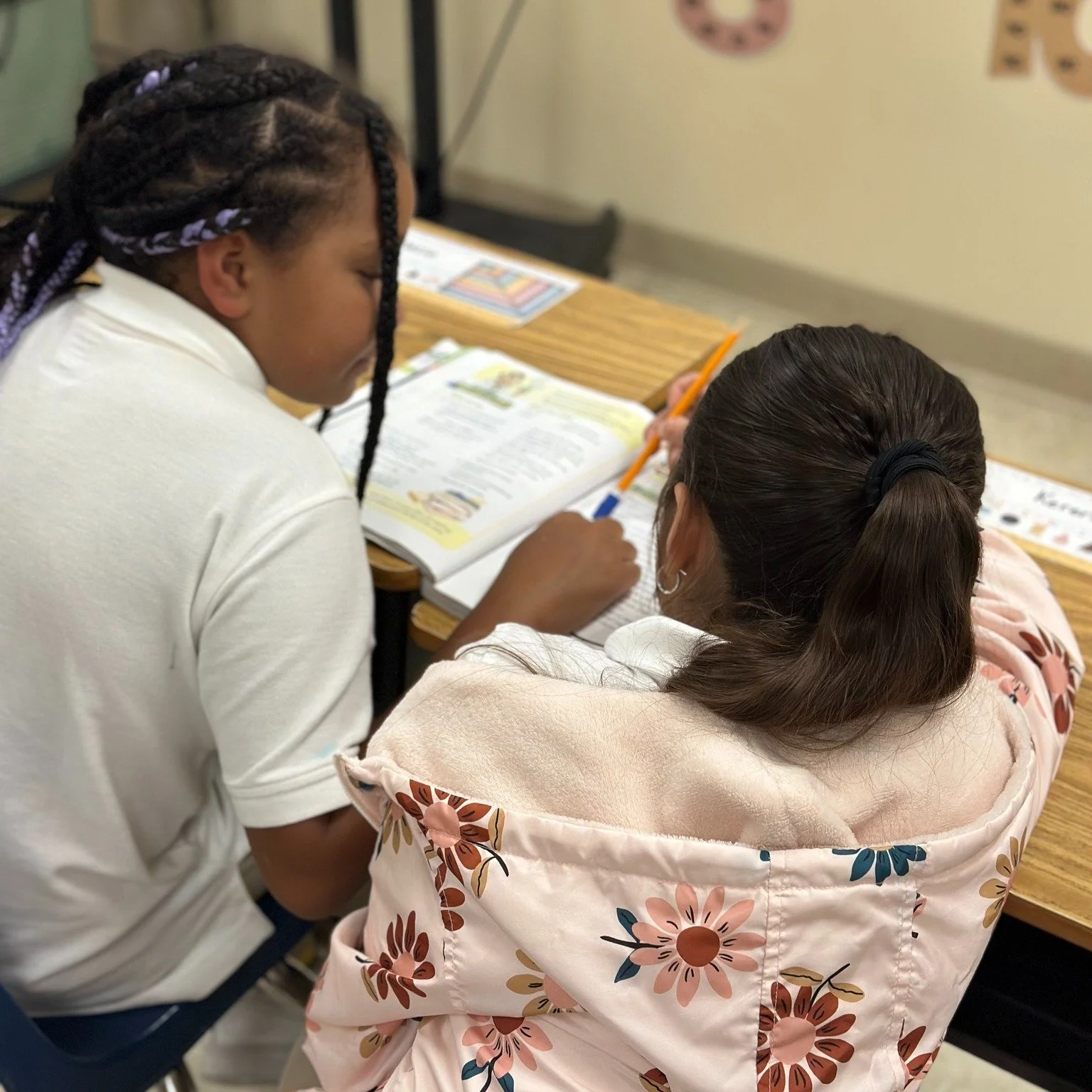 Two students sitting at a desk in a classroom, working together on an open textbook with colorful illustrations.