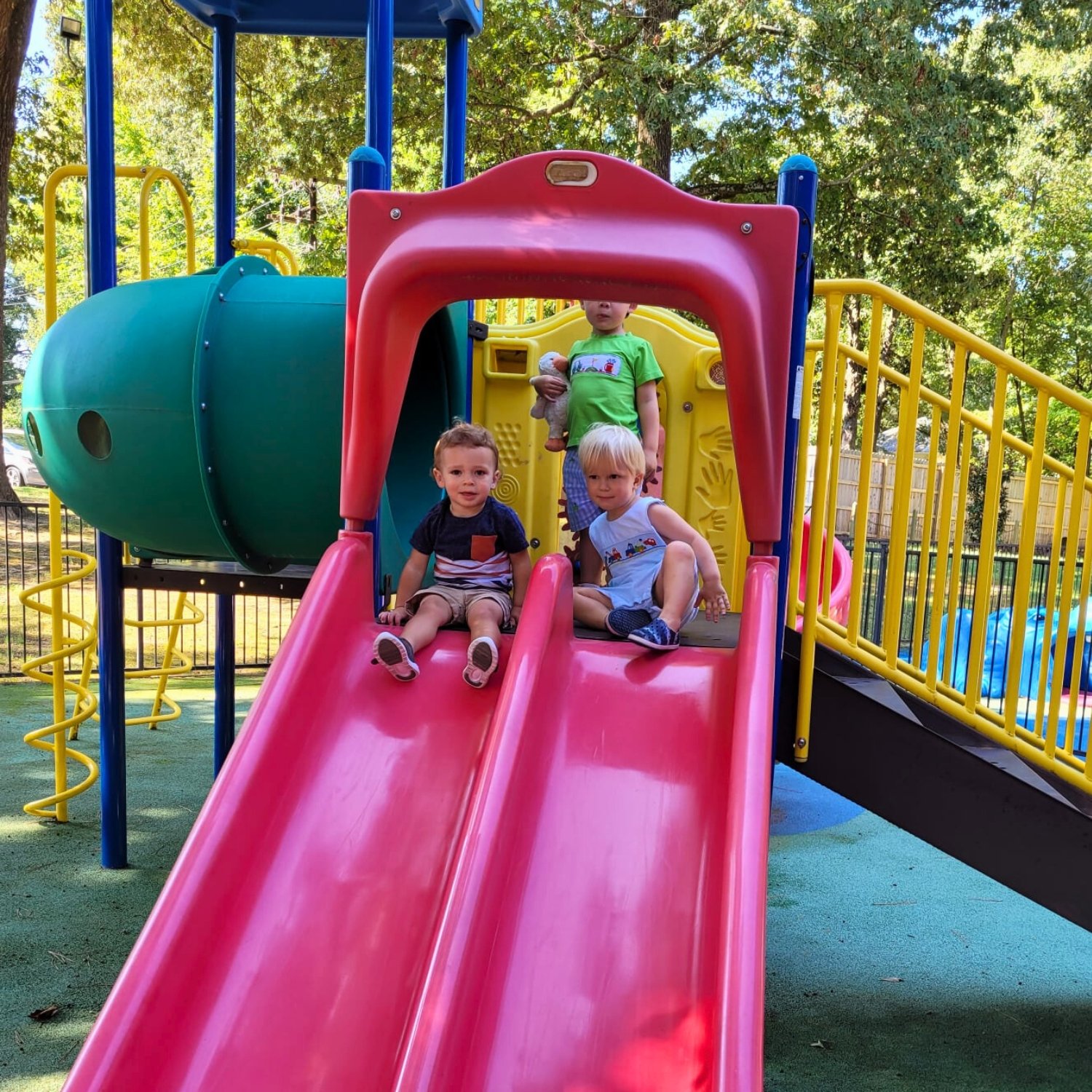 Three children playing on a colorful playground slide, with two sitting at the bottom and one standing behind them holding a stuffed animal.