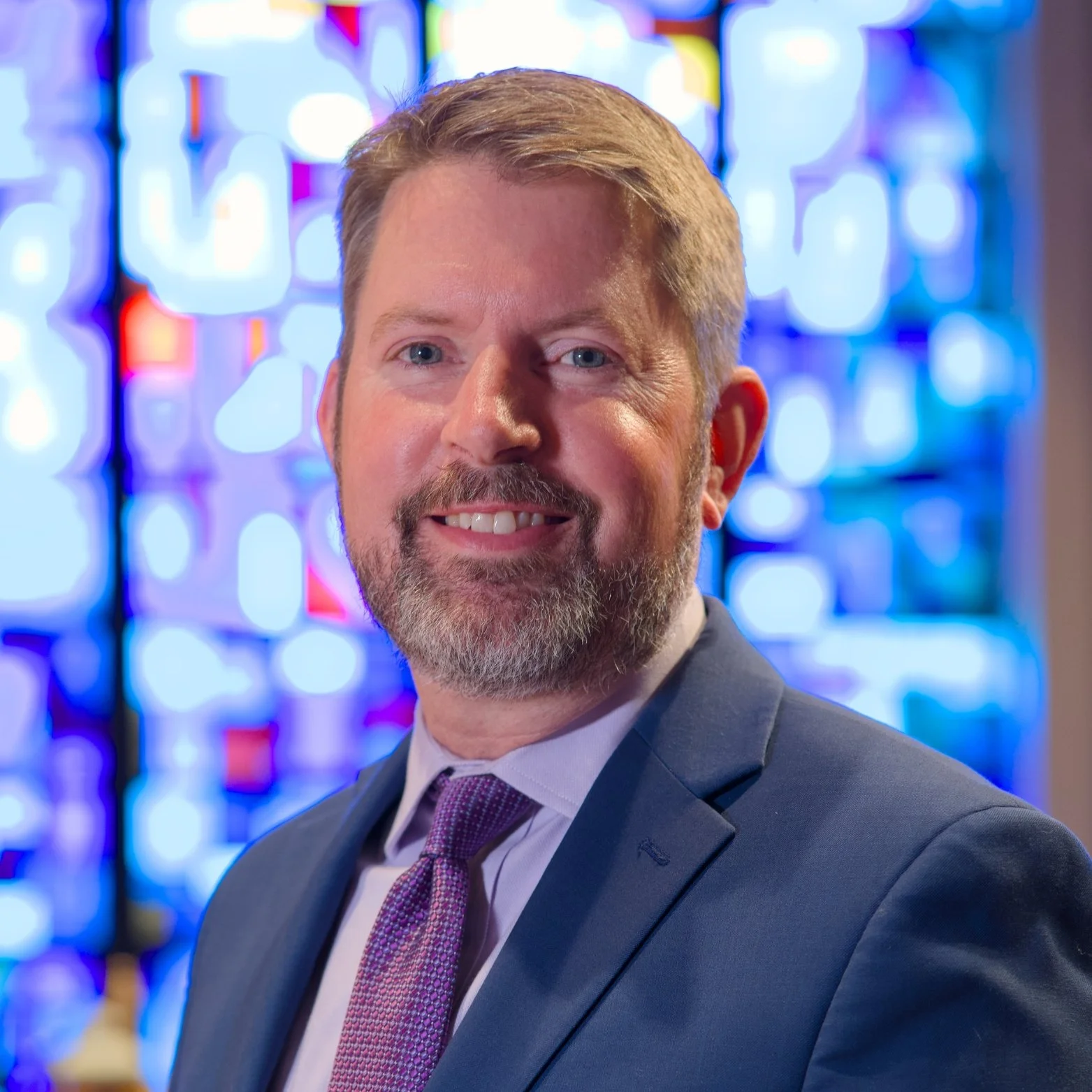 A man with short, light brown hair and a beard, wearing a dark suit, white shirt, and purple tie, smiling in front of colorful stained glass windows.