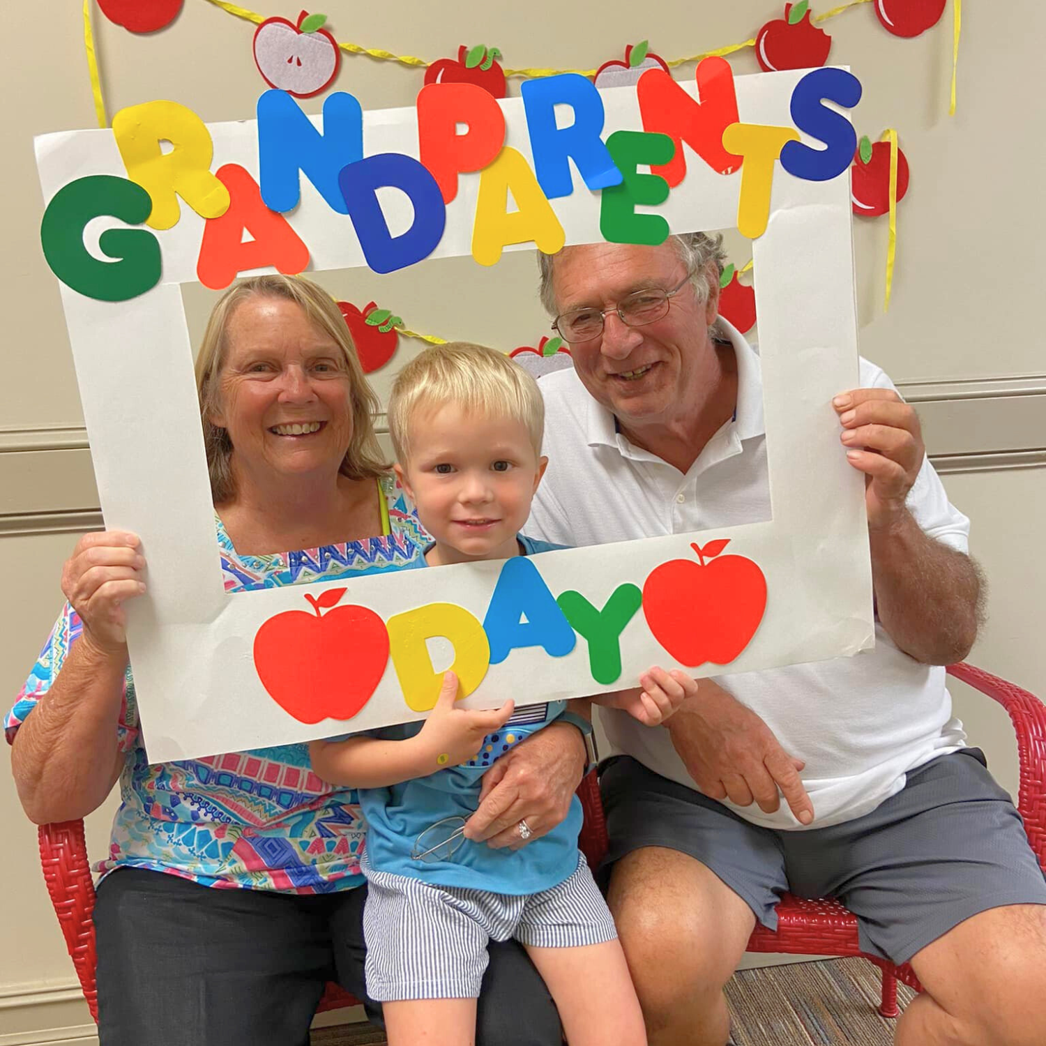 Three family members, two adults and one child, posing with a decorated frame that says 'Grandparents Day' with apple and letter decorations, celebrating grandparents' day.