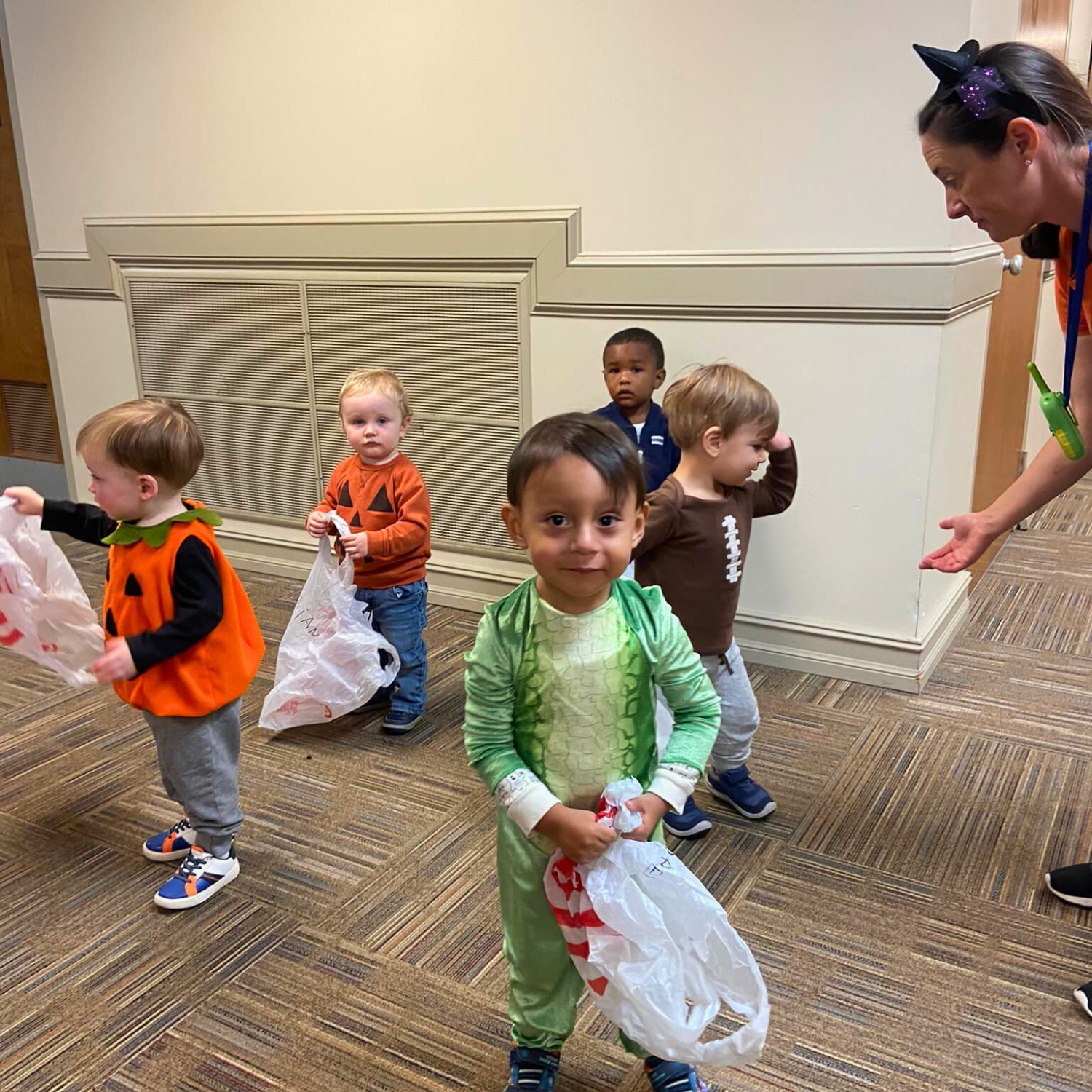 Group of young children in Halloween costumes standing in line indoors, some holding plastic bags for trick-or-treating, with a woman wearing a headband with a bow and nose instructing them.