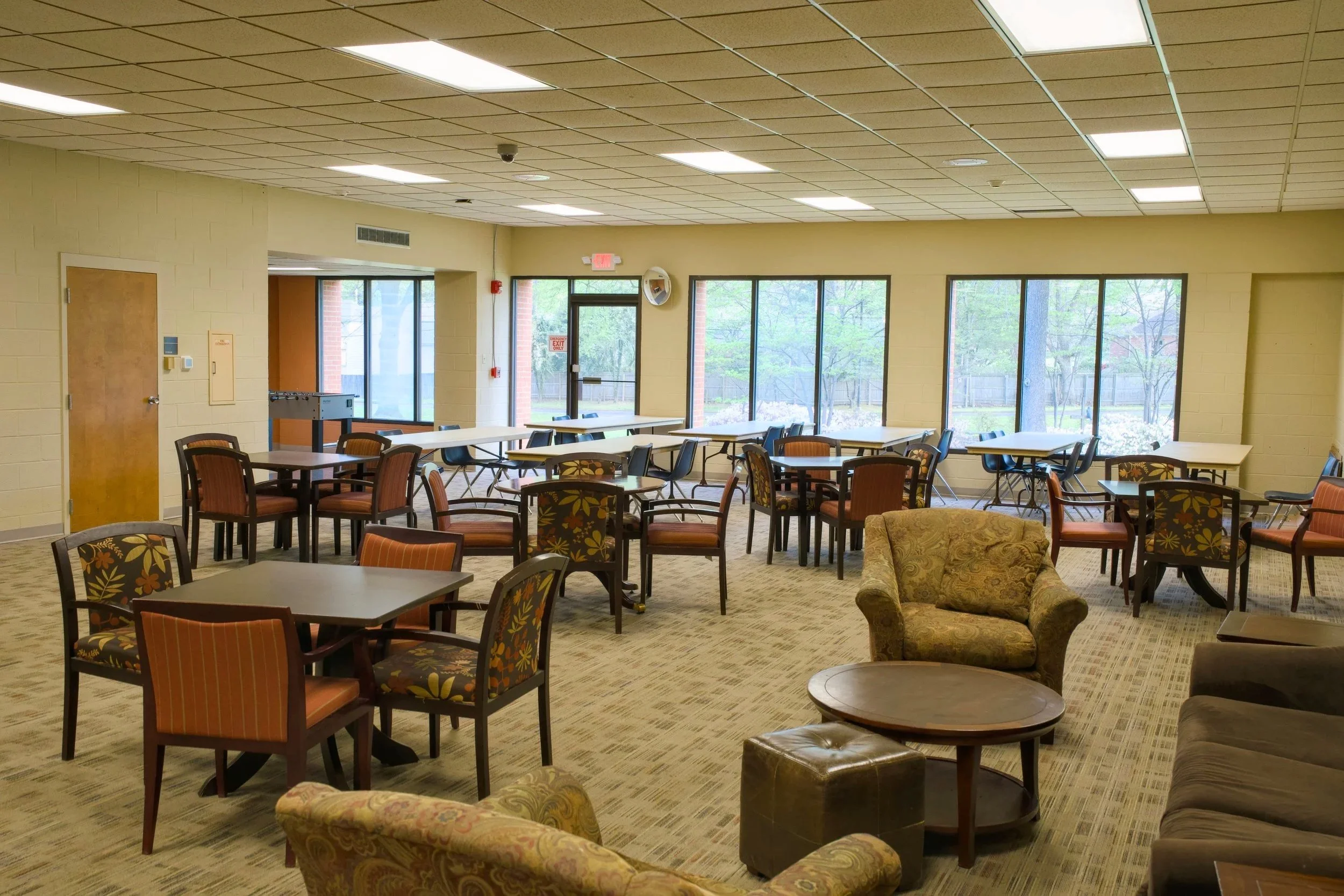 An empty communal or lounge area with tables, chairs, and a sofa, large windows showing greenery outside, and ceiling lights.
