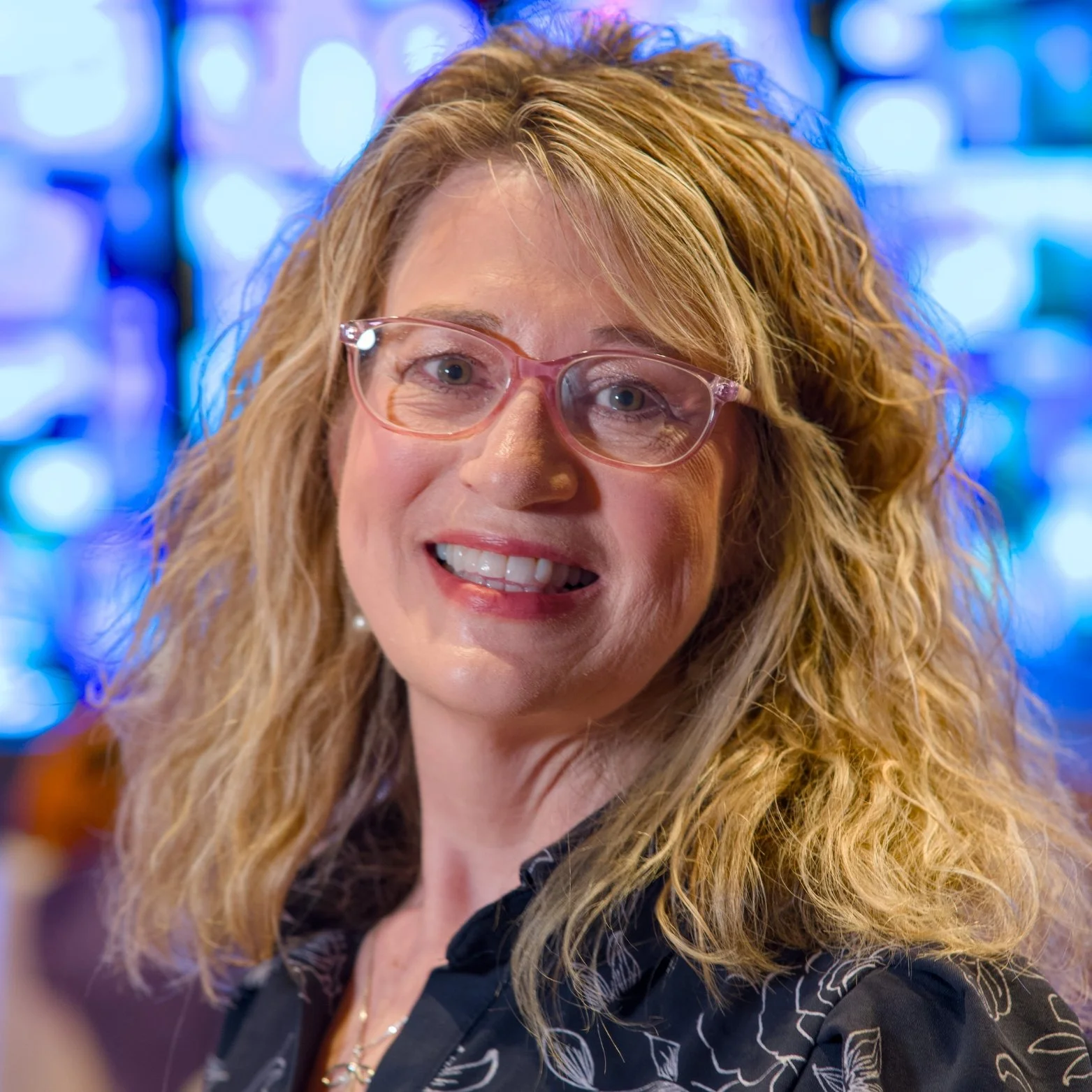 A woman with wavy blonde hair and pink glasses, smiling with her teeth showing, in a colorful, lit background.