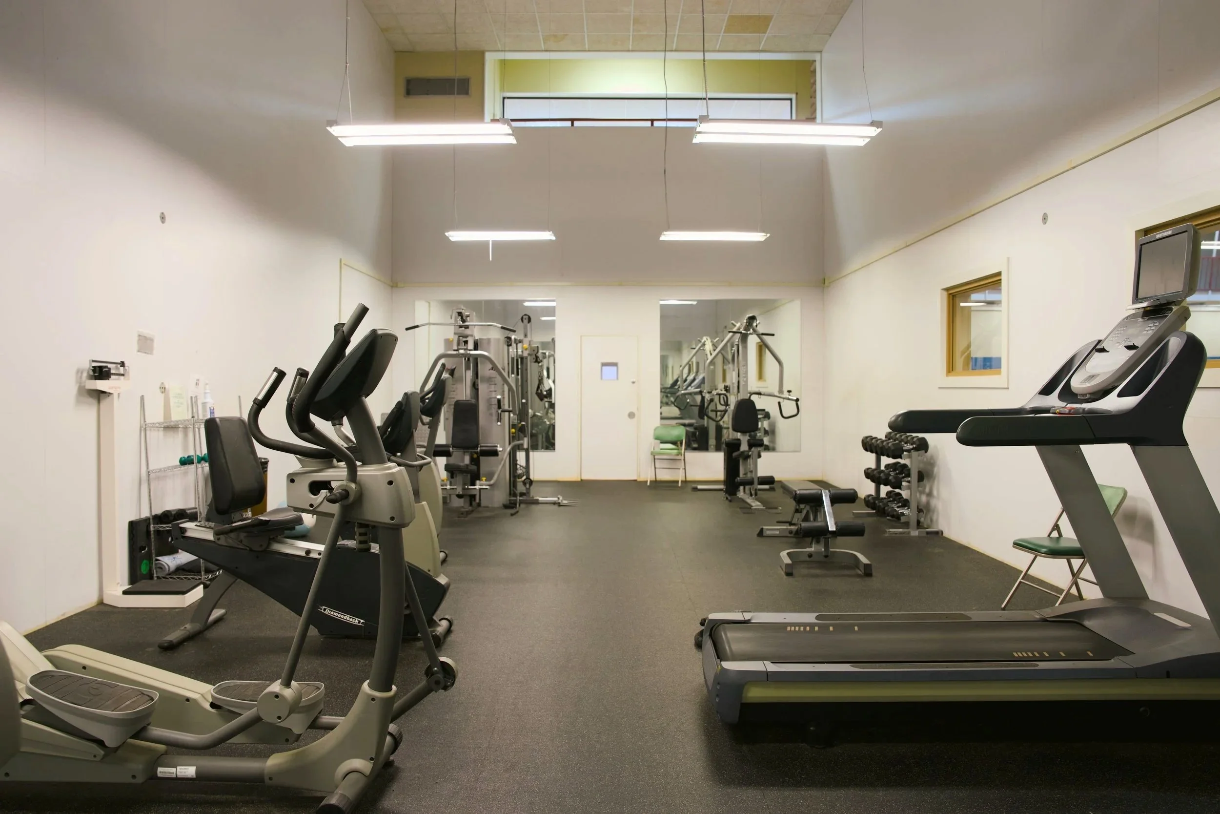 Empty gym with various exercise machines and weights, mirror on back wall, white walls and ceiling with fluorescent lighting.