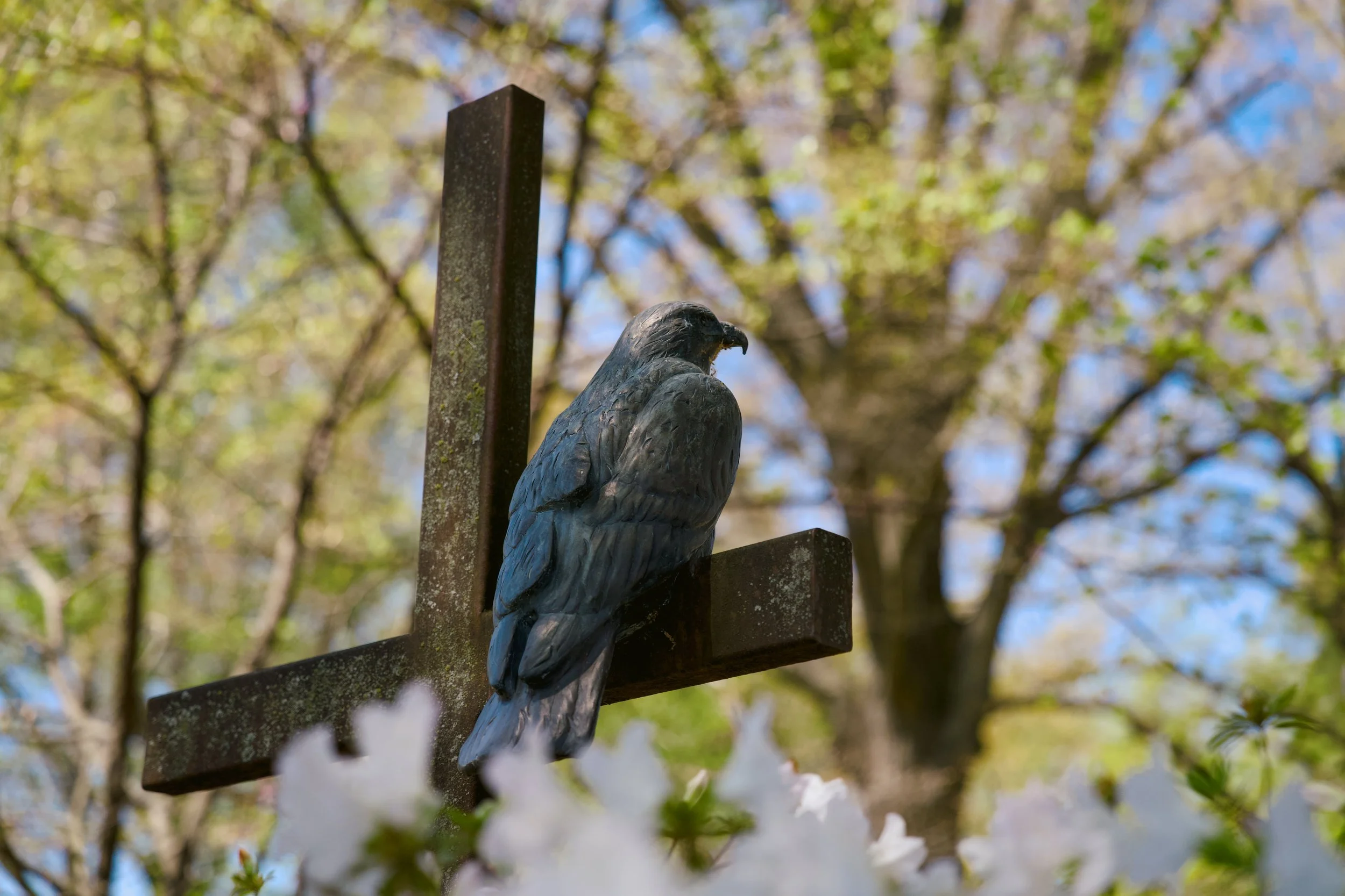 A close-up of a wooden cross with a metal eagle sculpture perched on it, set against a background of trees and blue sky.