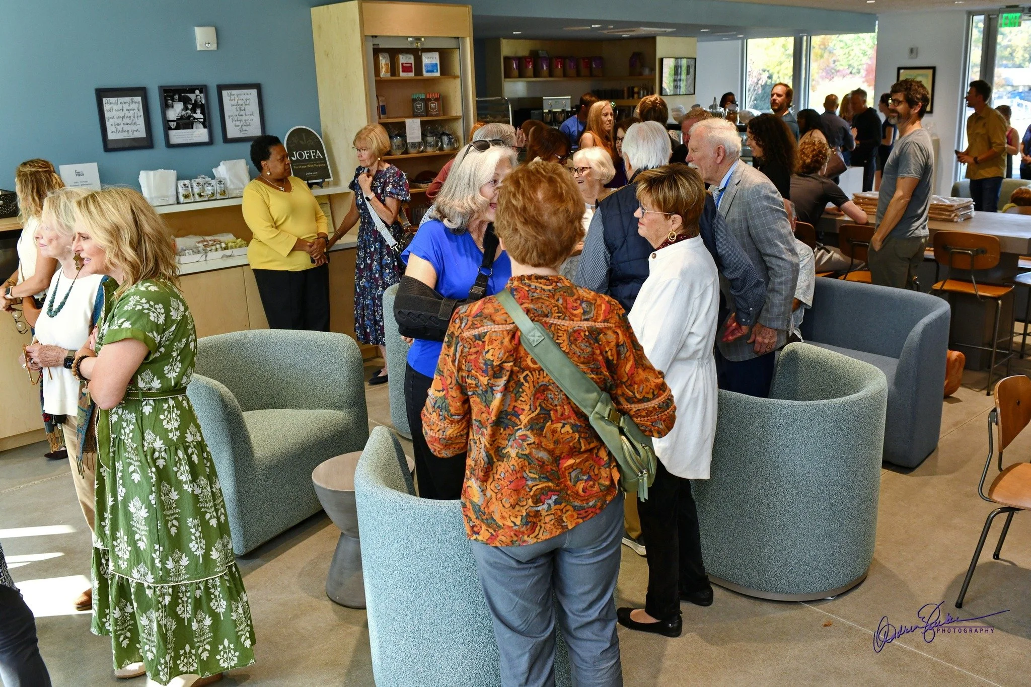 A group of people gathered inside a modern cafe, engaging in conversations, with some standing and others seated, in a well-lit setting with large windows and shelves with products in the background.