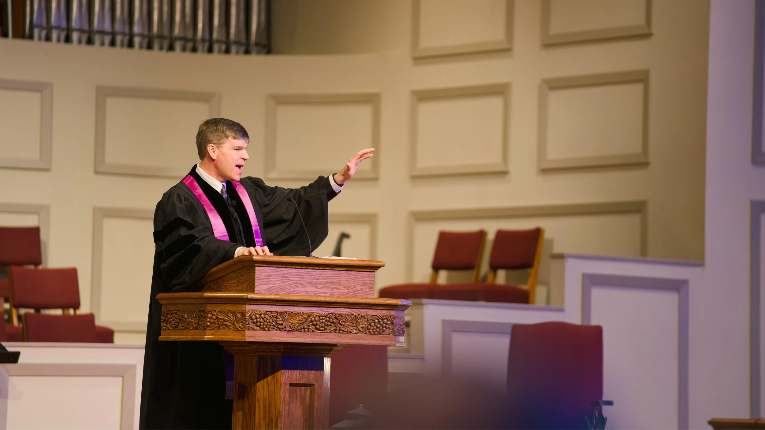 A man dressed in a black robe with purple accents is standing at a wooden pulpit, speaking and gesturing with his right hand inside a church or chapel with white paneled walls and empty red chairs in the background.
