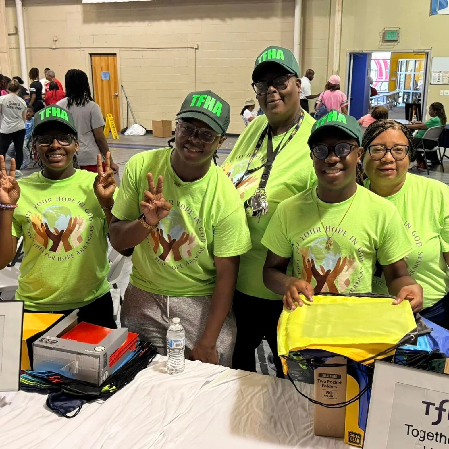 Group of five smiling volunteers wearing bright green T-shirts and caps with "TFHA" on them, standing behind a table at a community event, making peace signs. The table has supplies including water bottles, folders, and yellow drawstring bags, inside a large indoor venue.