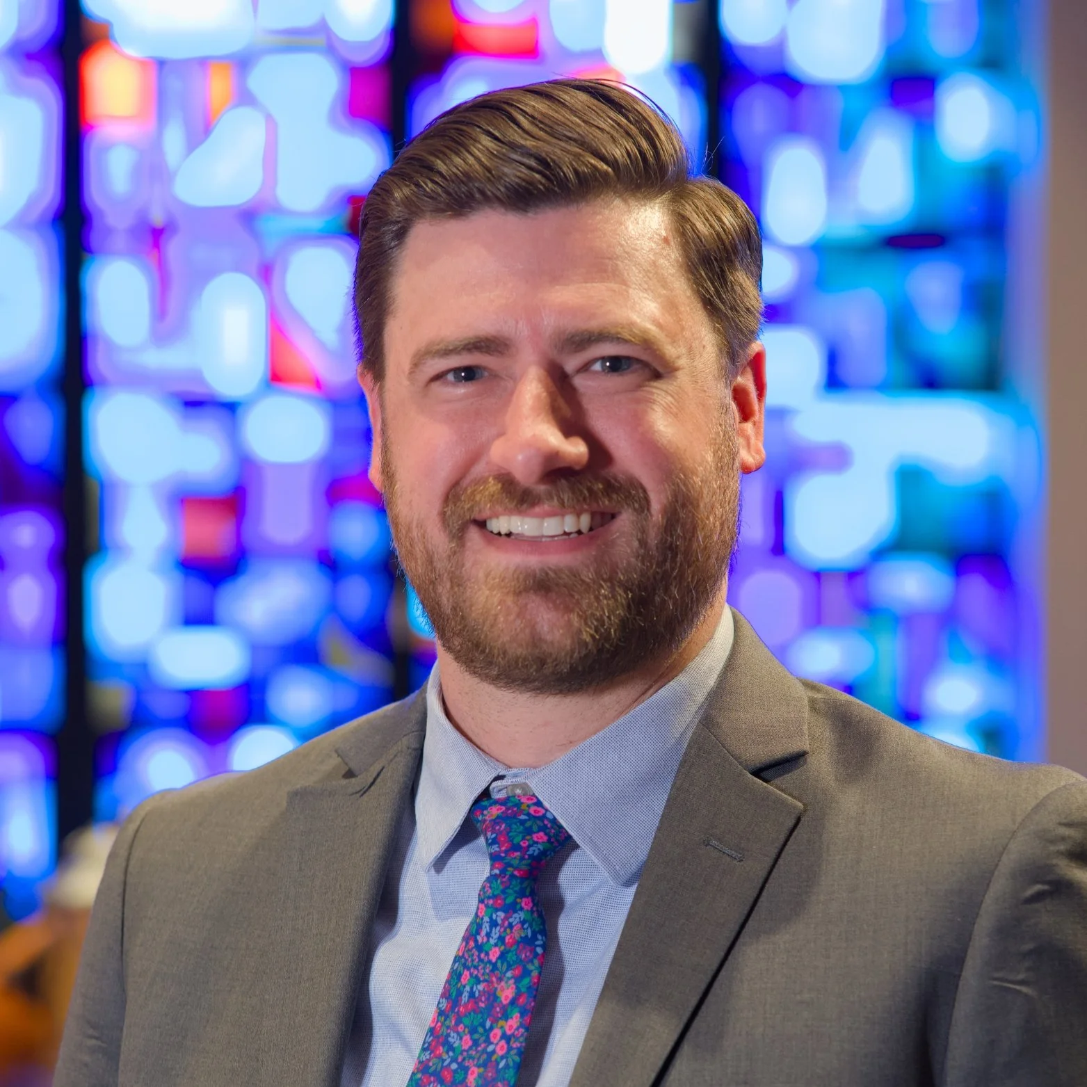 A smiling man in a gray suit and a colorful floral tie posing indoors with colorful stained glass windows in the background.