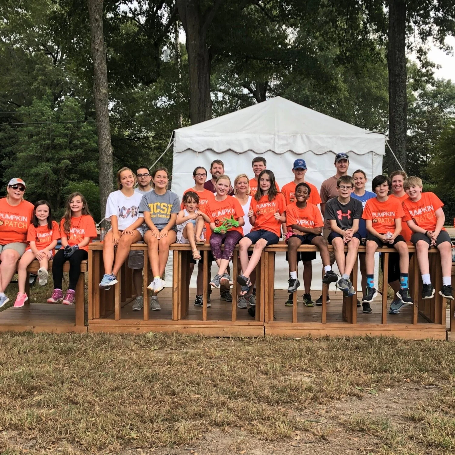Group of children and adults sitting on a wooden stage outdoors with trees and a white tent in the background, many wearing orange shirts that say 'Pumpkin Patch'.