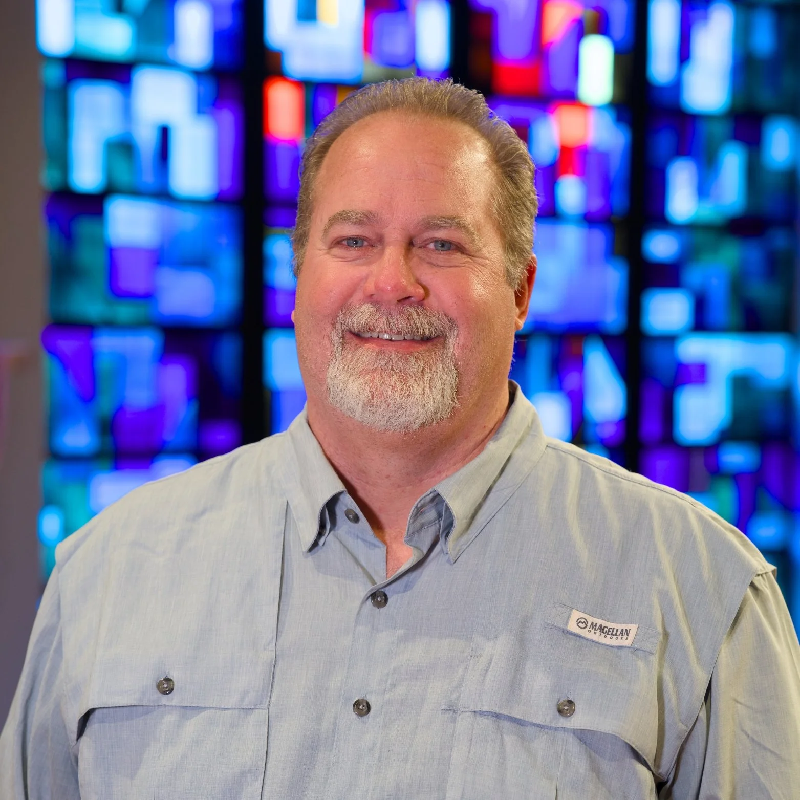 A smiling man with a beard and mustache, wearing a light-colored button-up shirt, standing in front of a colorful abstract background made of illuminated panels.