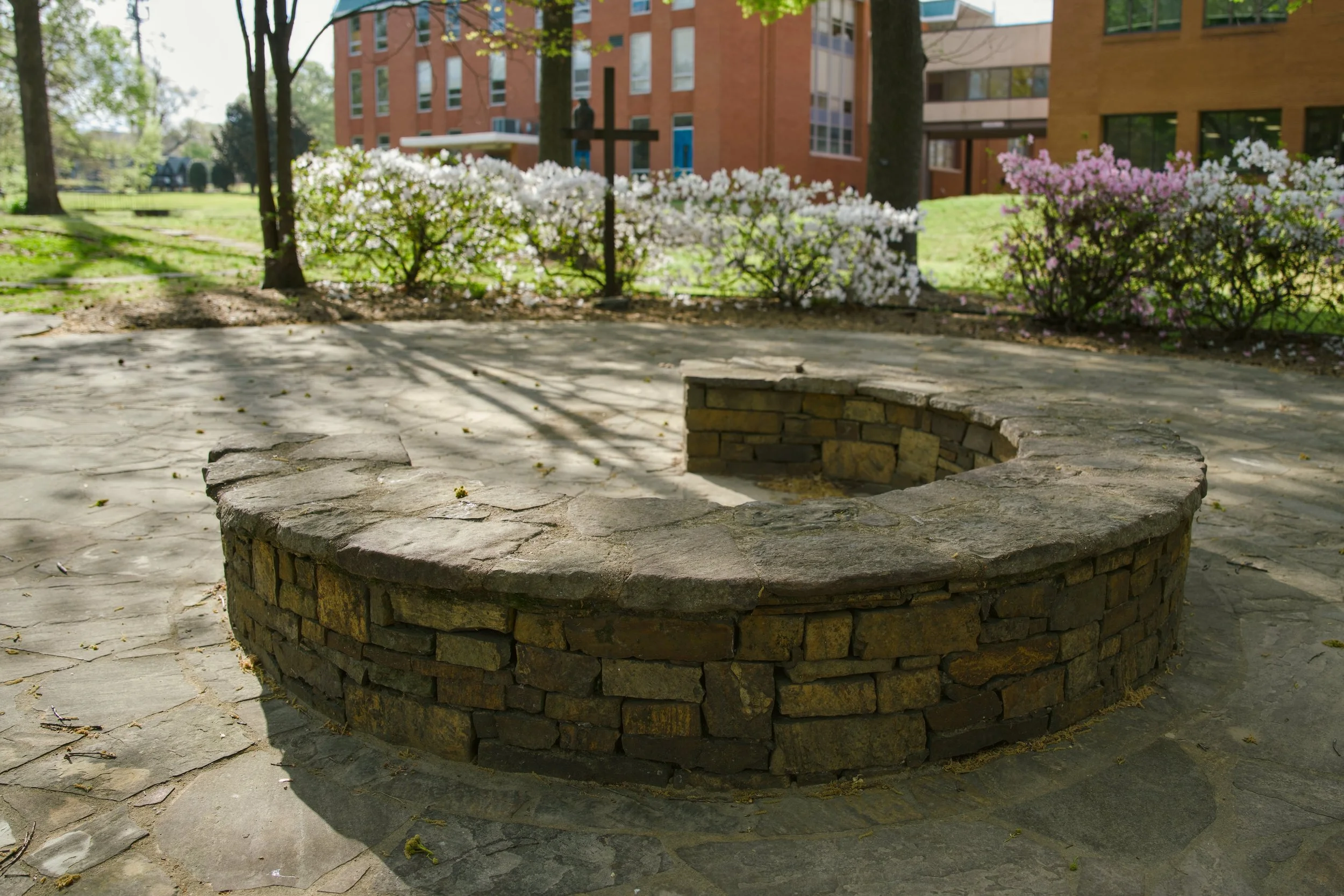 A circular stone fire pit with a brick inner wall, situated on a concrete patio surrounded by blooming bushes and trees in a park-like area, with buildings in the background.