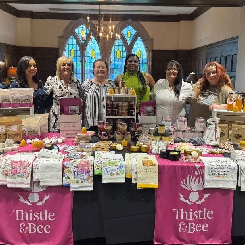 Six women standing behind a table displaying handmade products at a Thistle & Bee market booth inside a church with stained glass windows.