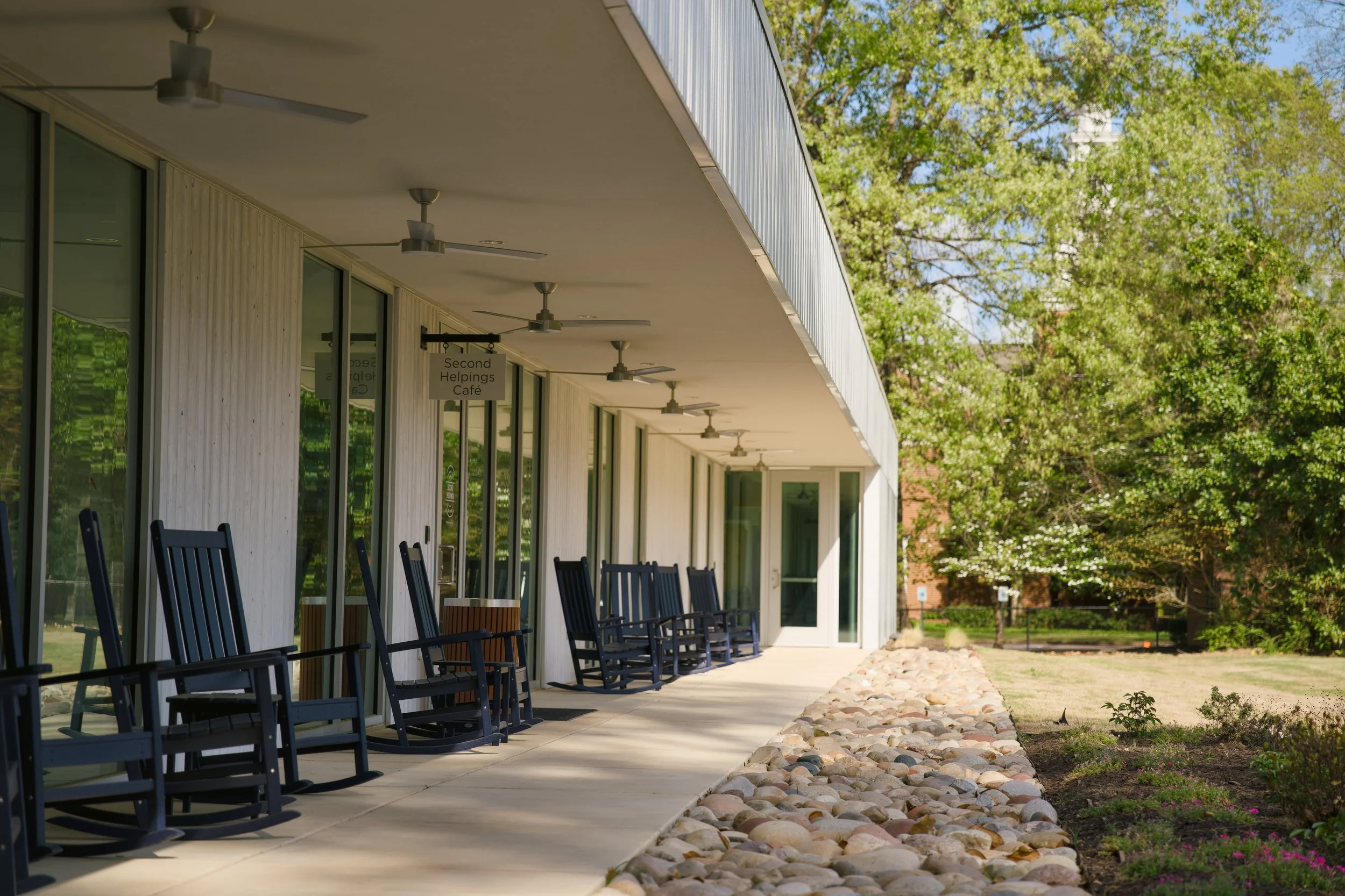Exterior view of a building with large glass windows and a covered walkway with several black rocking chairs. There's a sign that reads 'Second Helpings Café' above the walkway. The building is surrounded by a garden with rocks and greenery, and trees in the background.