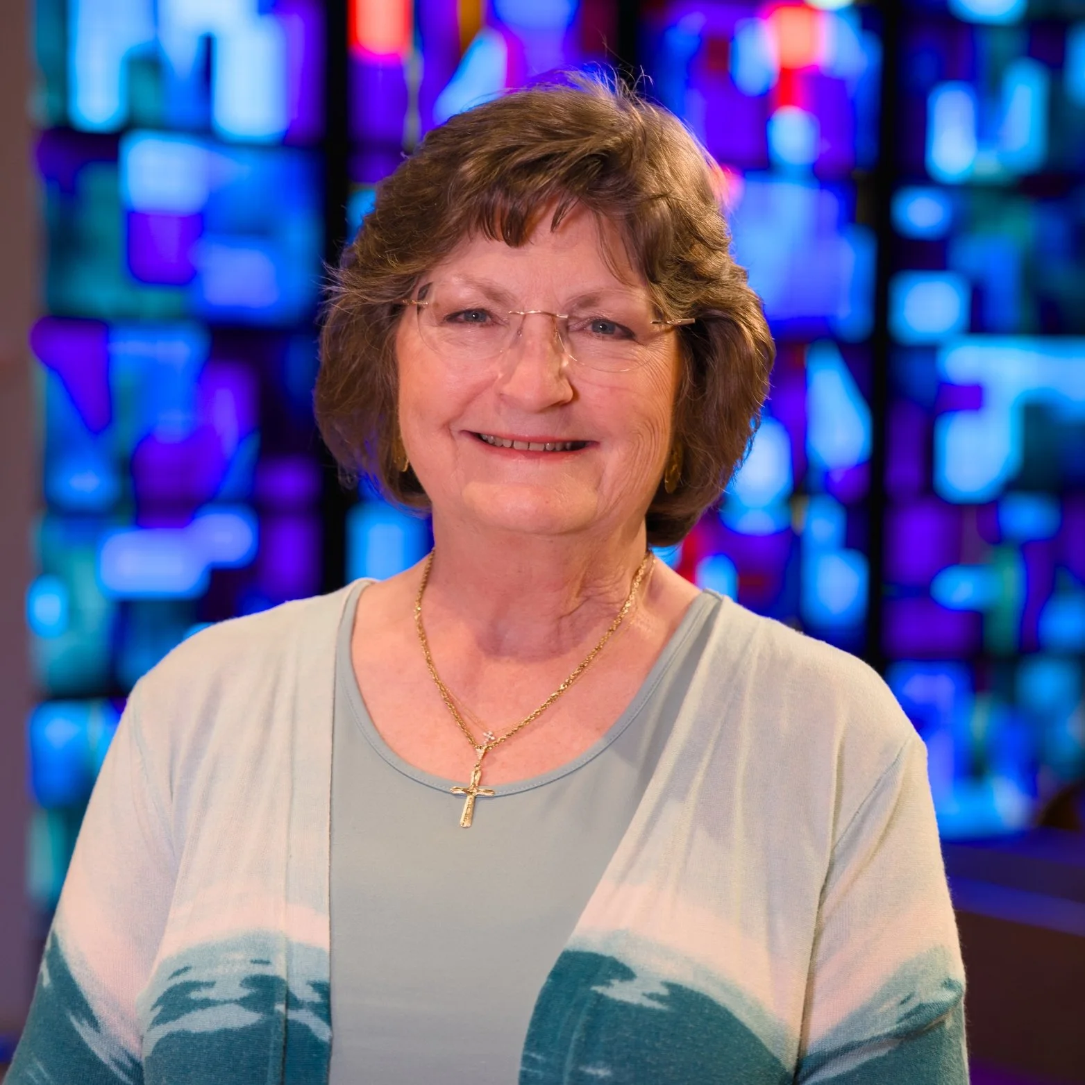 Portrait of an elderly woman with glasses, wearing a light-colored top and a gold cross necklace, smiling in front of a colorful stained glass background.