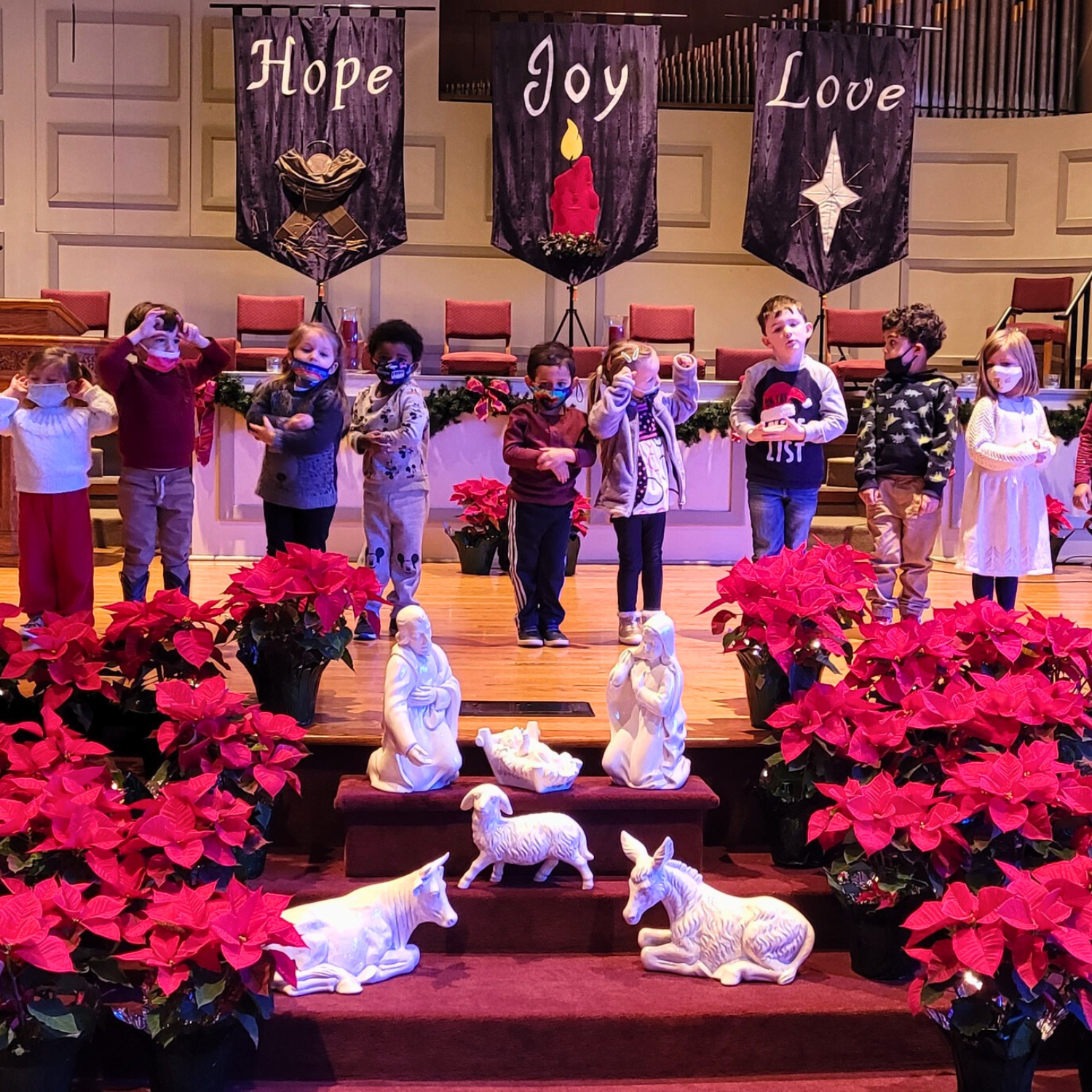Children standing on a stage with Christmas decorations, poinsettias, and nativity scene statues, with banners reading Hope, Joy, Love in the background.