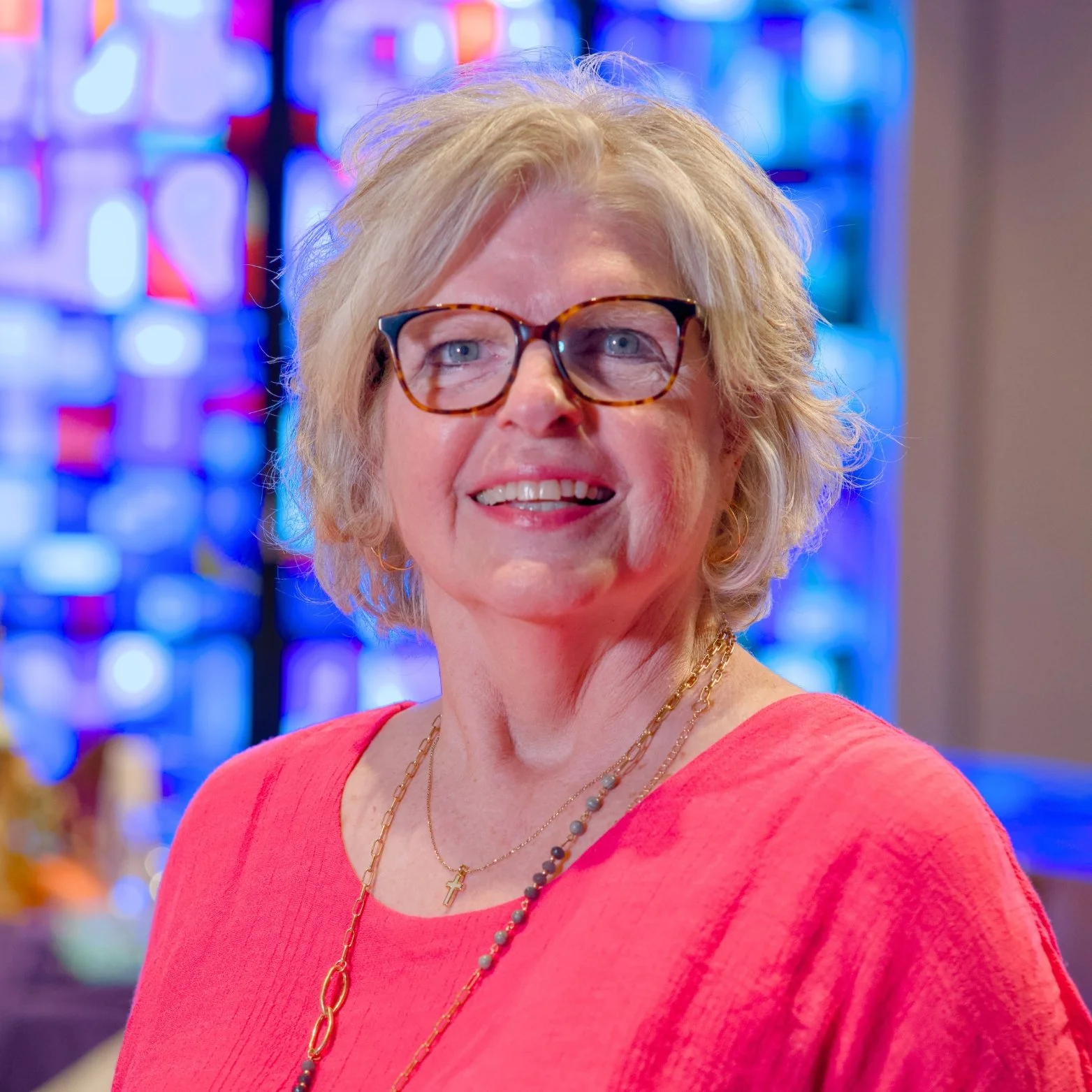 A woman with short, curly blonde hair and glasses, smiling, wearing a red top, with colorful stained glass window in the background.