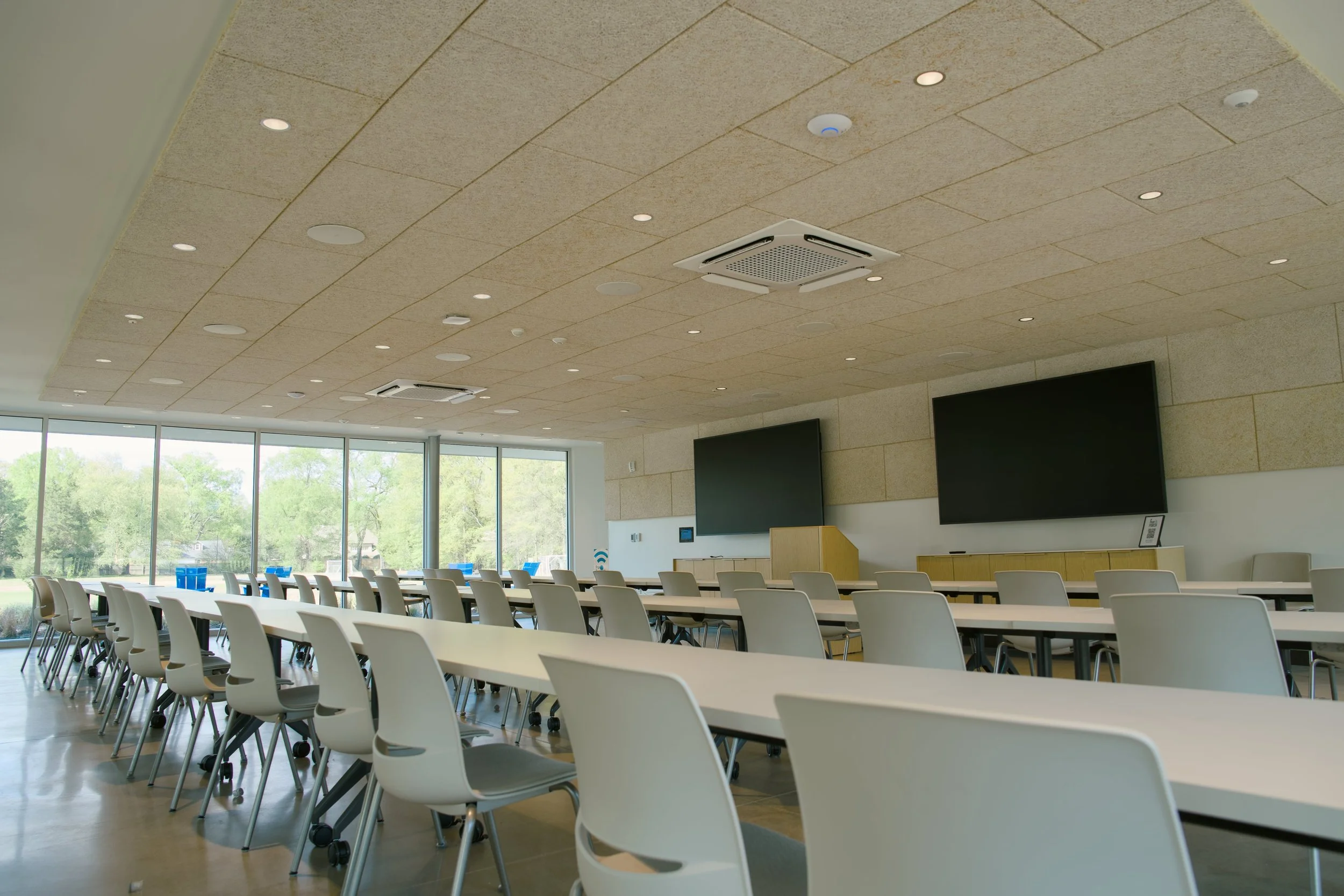 Modern conference room with rows of white chairs and long tables, two large black screens on the wall, large windows showing green trees, and ceiling with recessed lights and ventilation.