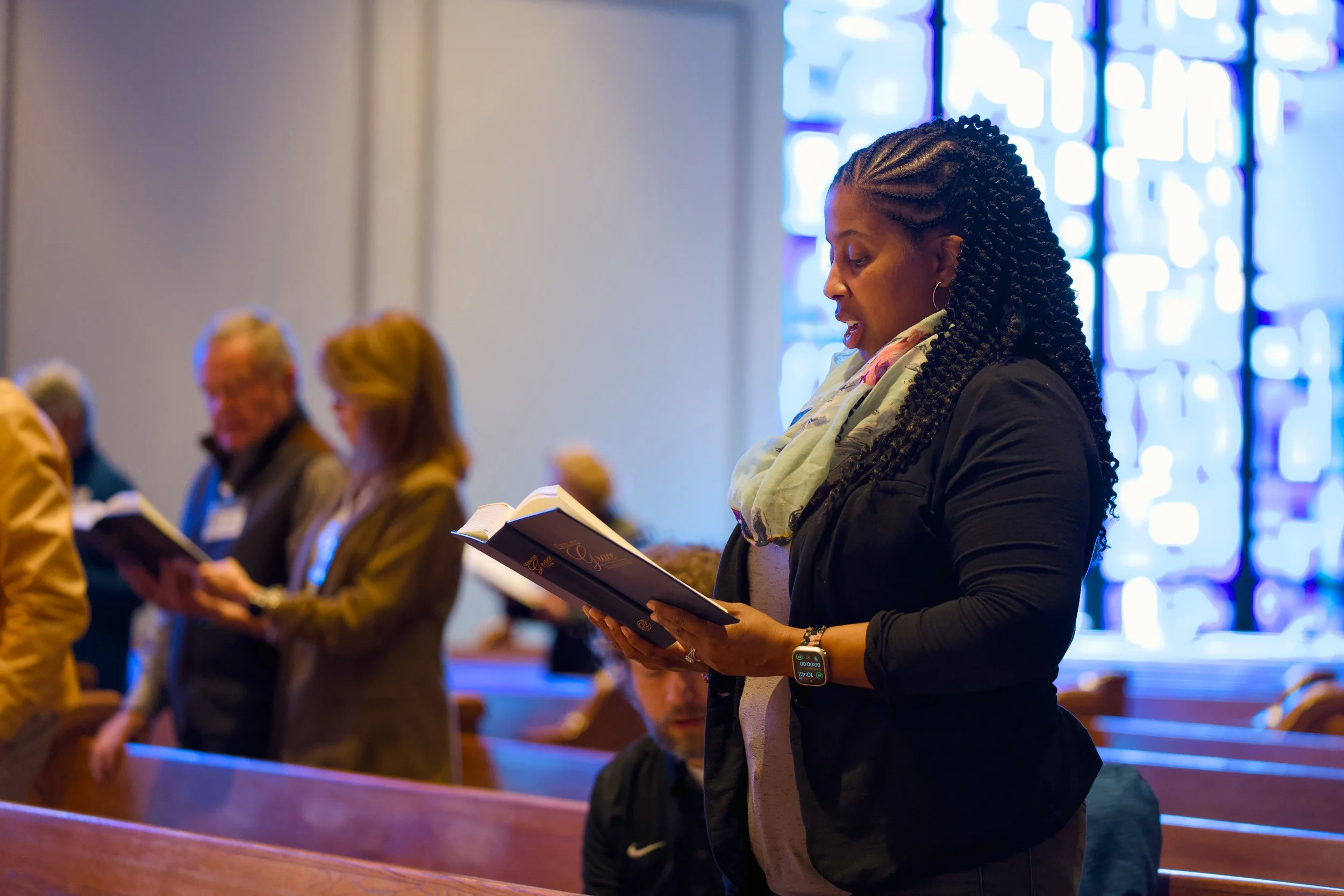 A woman with braided hair and a scarf standing in a church pew, reading from a book during a church service.