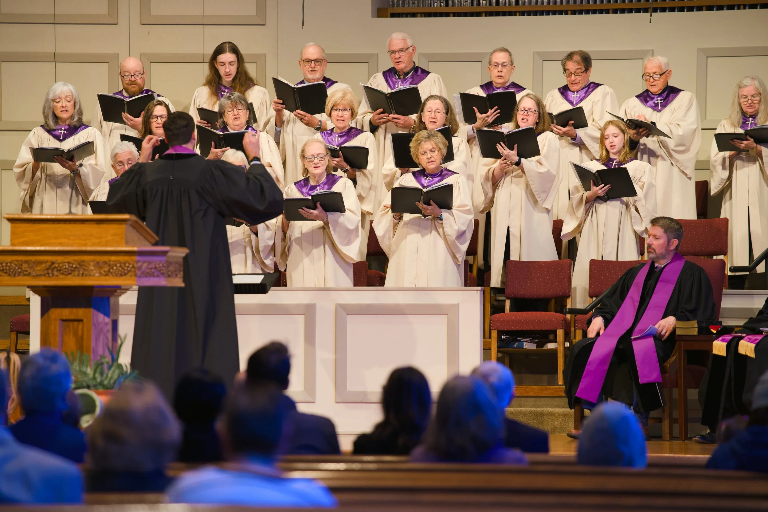 A church choir performing on stage, directed by a conductor, with an audience watching.
