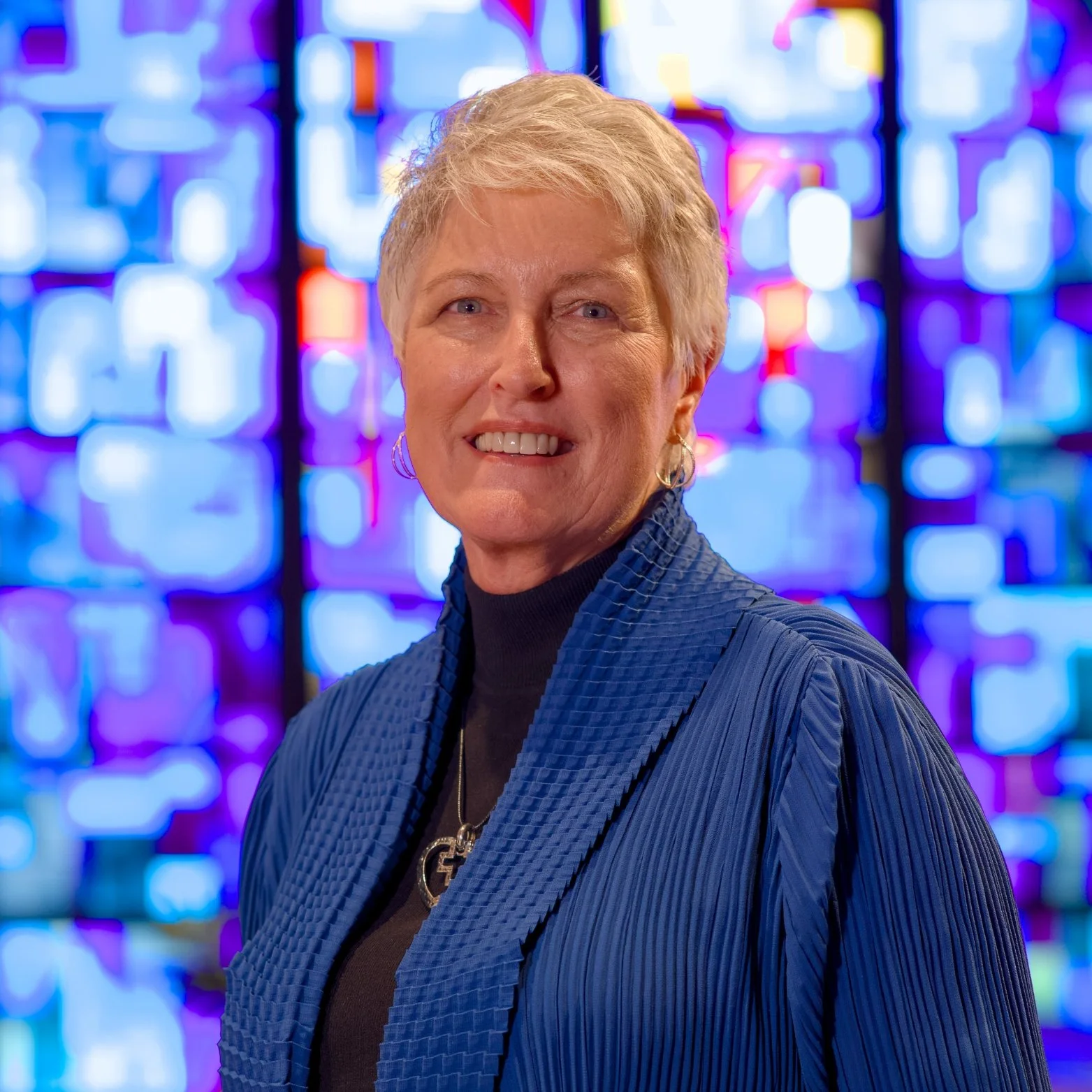 A smiling woman with short blonde hair, wearing a blue textured jacket, black turtleneck, and a cross necklace, standing in front of a colorful, stained glass window.