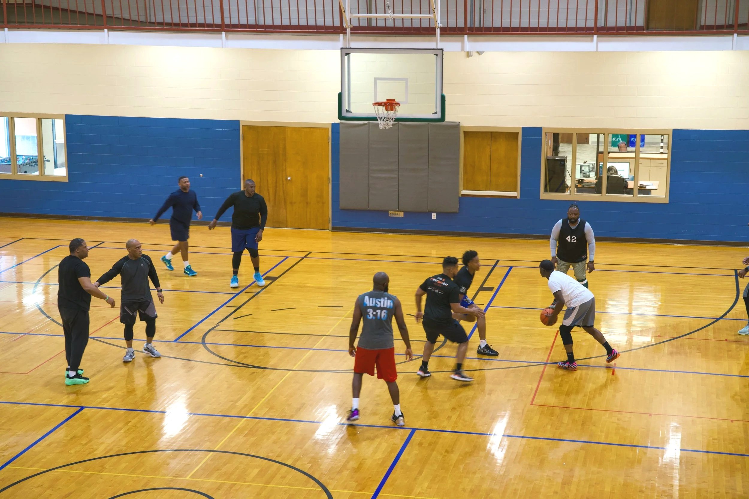 A group of men playing basketball on an indoor court with wooden flooring, blue walls, and a basketball hoop.