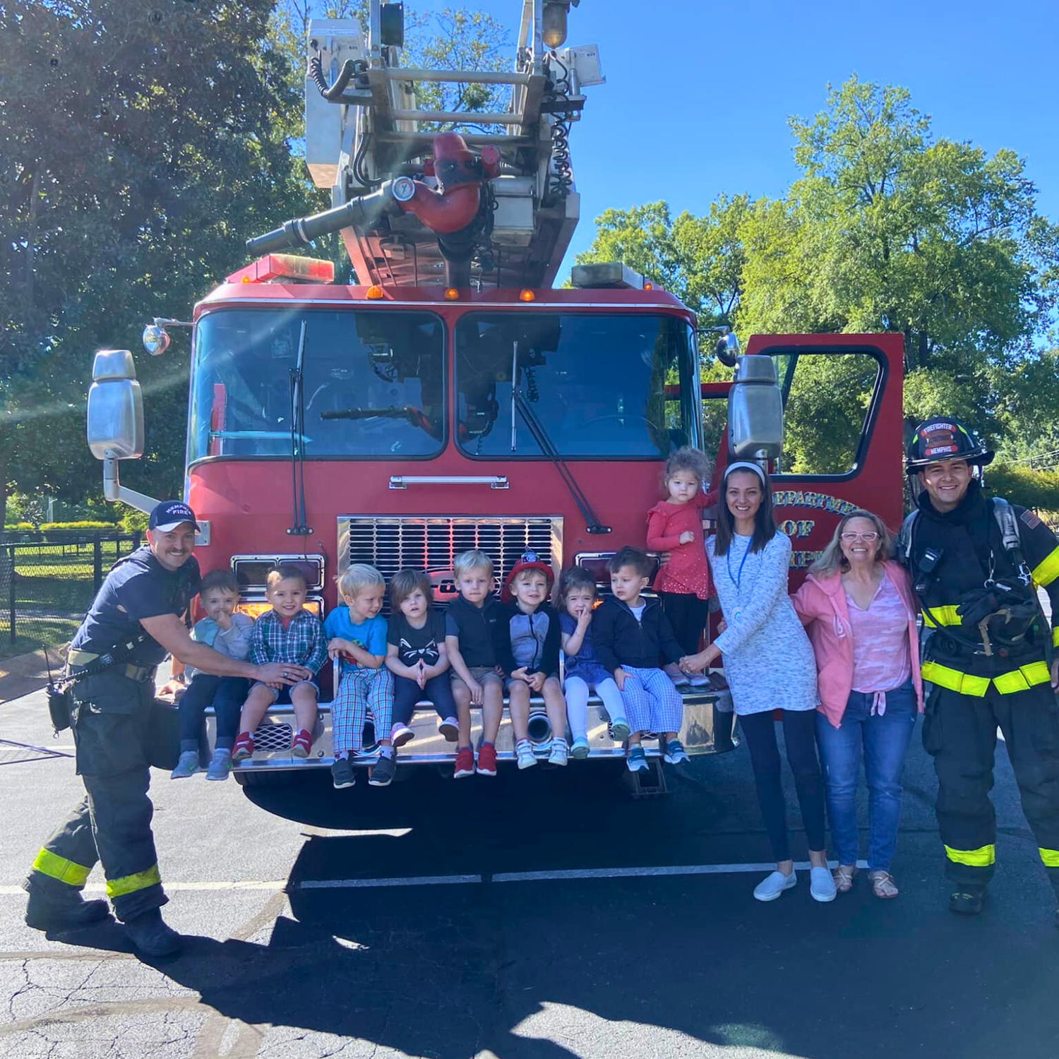 A group of young children and adults, including firefighters, sit and stand on the front of a red fire truck during a daytime event. The children are sitting on the truck's bumper, and some are holding hands with the adults. The firefighters are in uniform, smiling, and posing with the group in an outdoor setting with green trees and a clear blue sky.