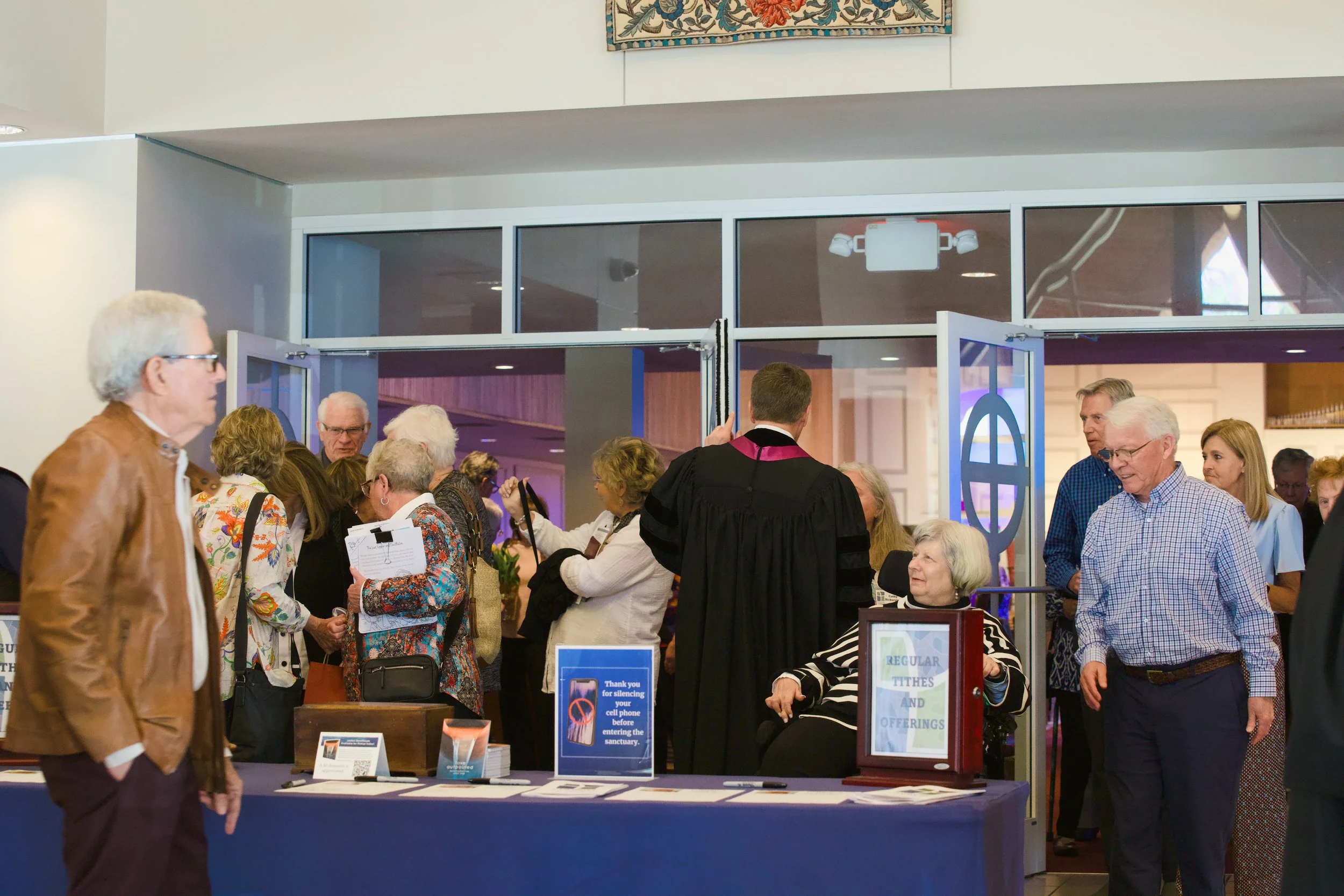 Group of elderly people and a man in judicial robes gathered near a registration table at a church or community event.