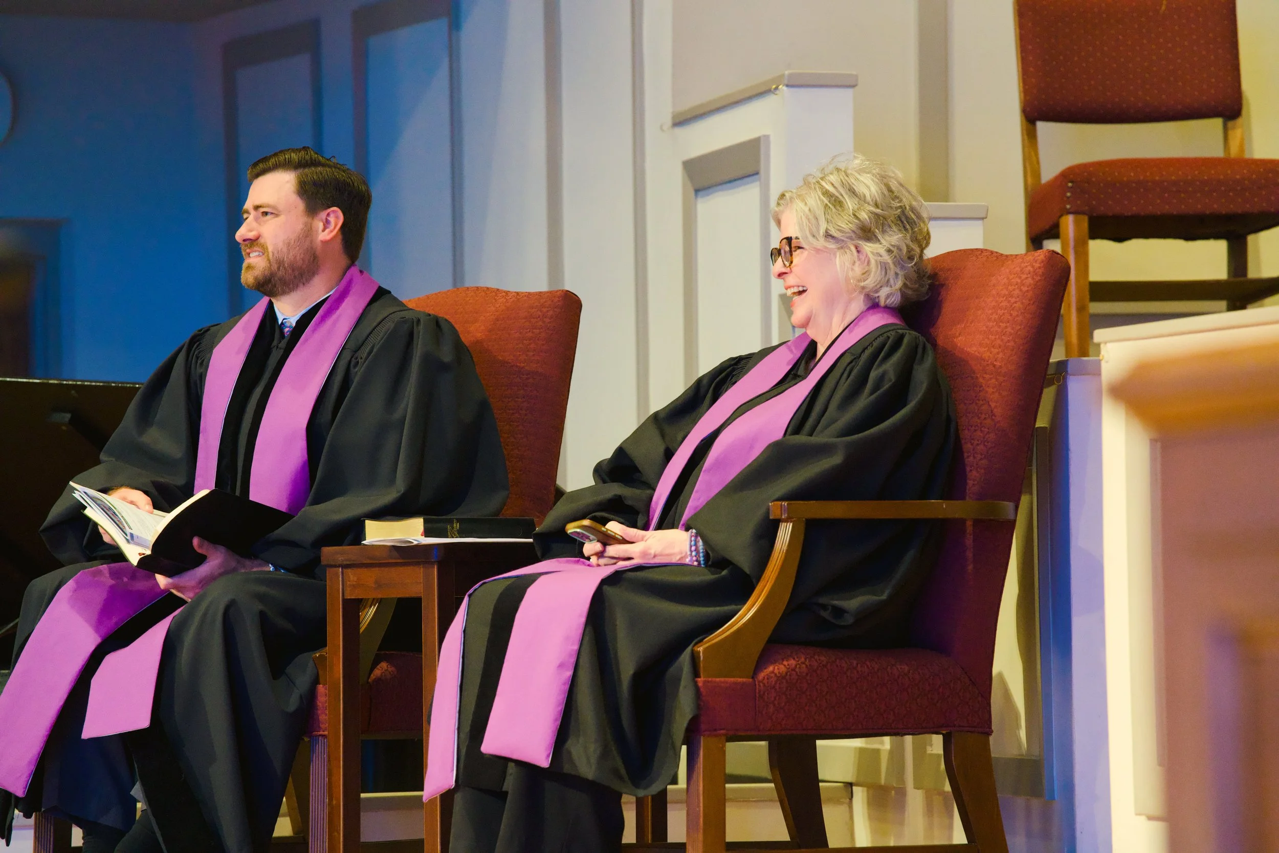 Two people wearing graduation robes with purple stoles, seated in chairs on a stage, smiling and laughing, one holding a booklet and the other holding a phone.