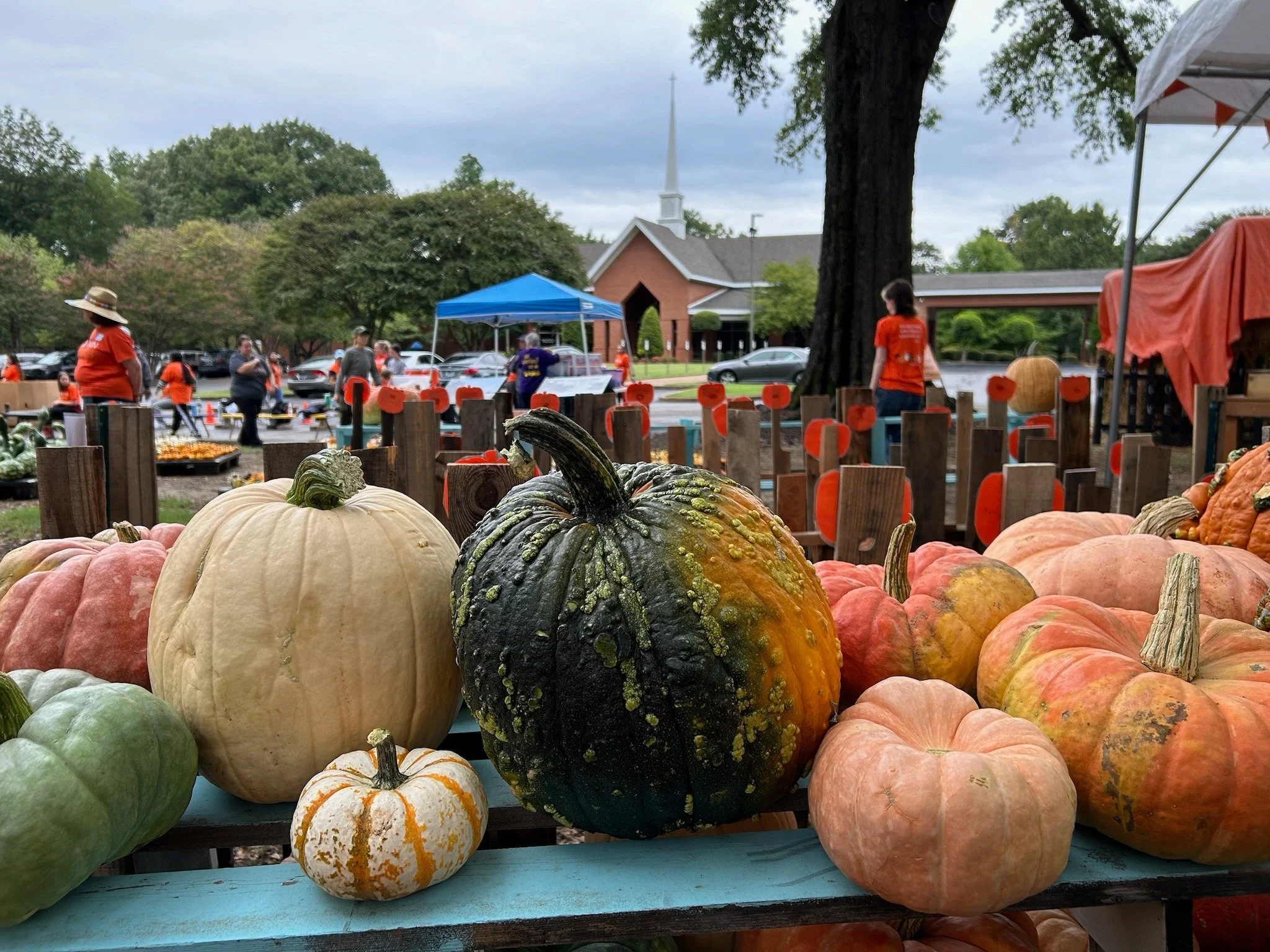 Colorful pumpkins in the foreground at an outdoor market, with people shopping and a church in the background on a cloudy day.
