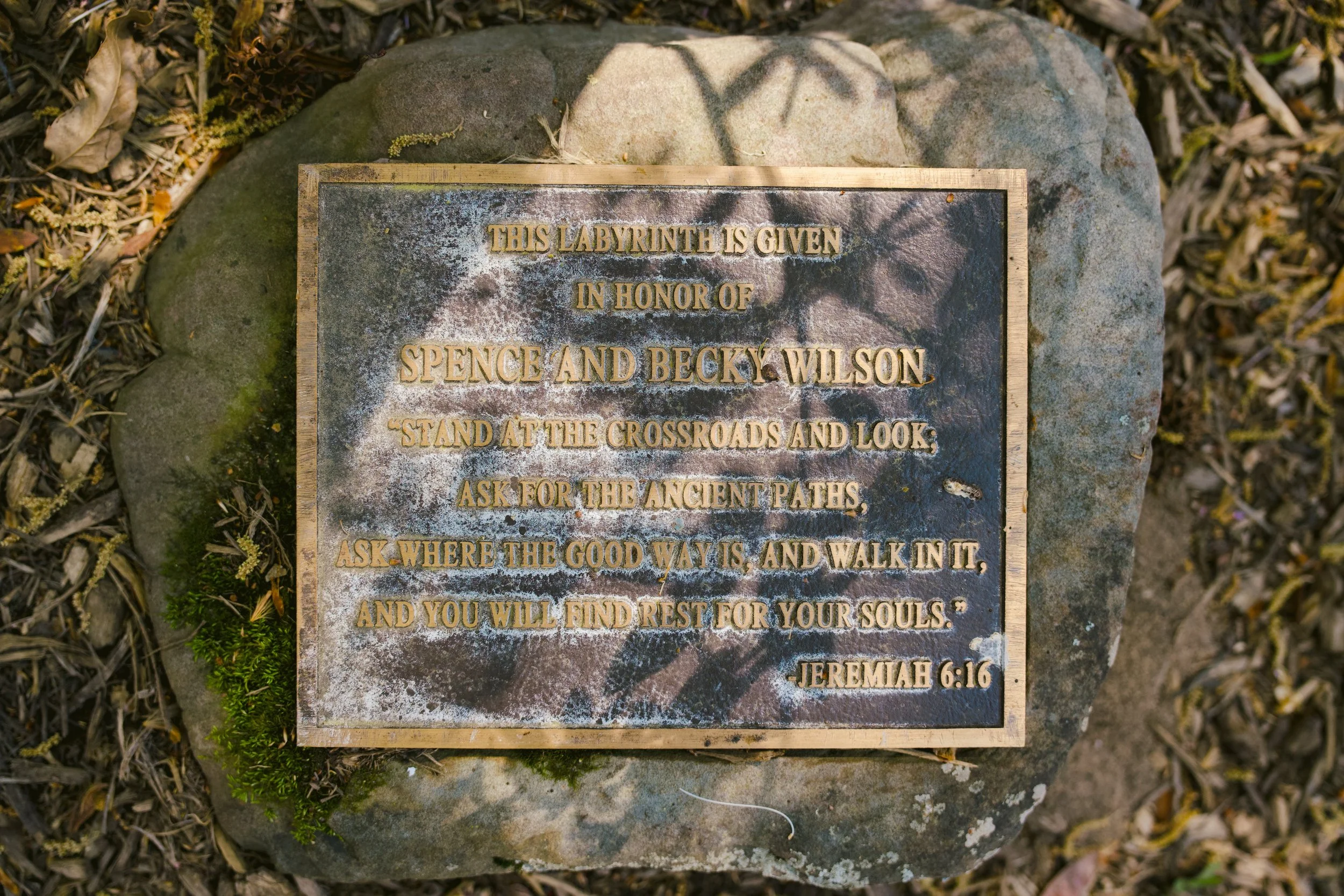 A bronze plaque mounted on a rock with an etched quote from Jeremiah 6:16, surrounded by moss, leaves, and small plants.