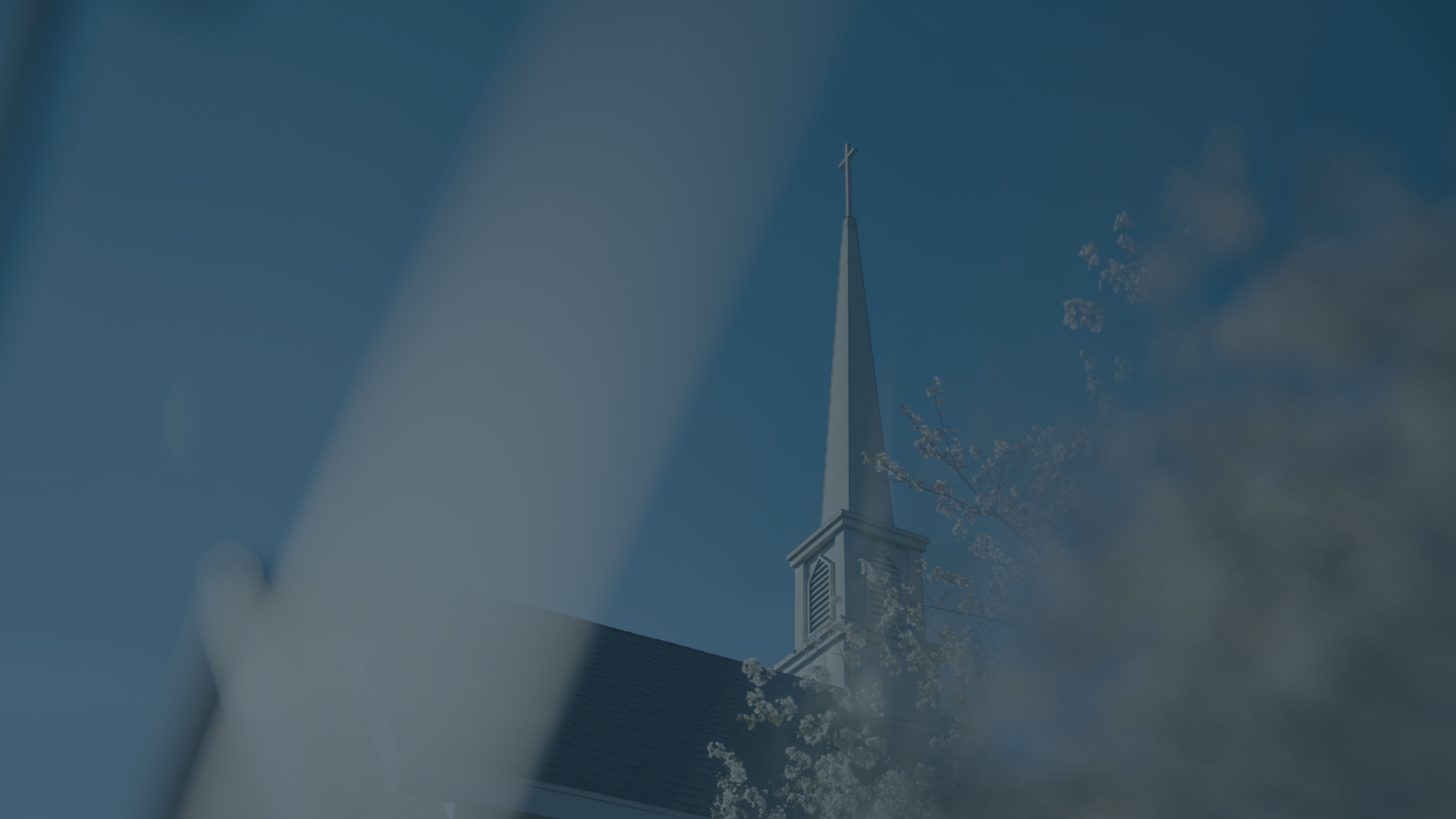 A church steeple with a cross on top, framed by blurred branches and clouds in a blue sky.