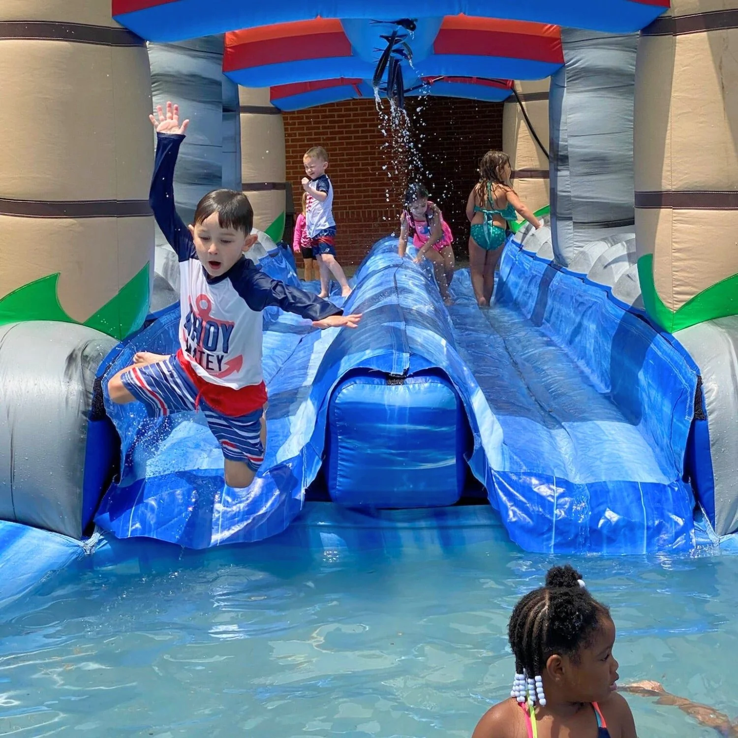 Children playing on an inflatable water slide in a pool, with some splashing and running, at a sunny day. A girl with braided hair is in the pool, and a boy with striped swim shorts is jumping off the slide.