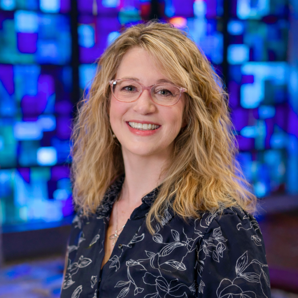 A woman with curly blonde hair and glasses smiling in front of a colorful, abstract background.