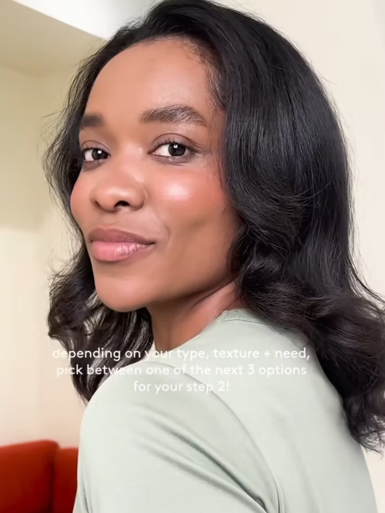 Close-up of a woman with medium skin tone, black wavy hair, and a slight smile, wearing a light-colored top.