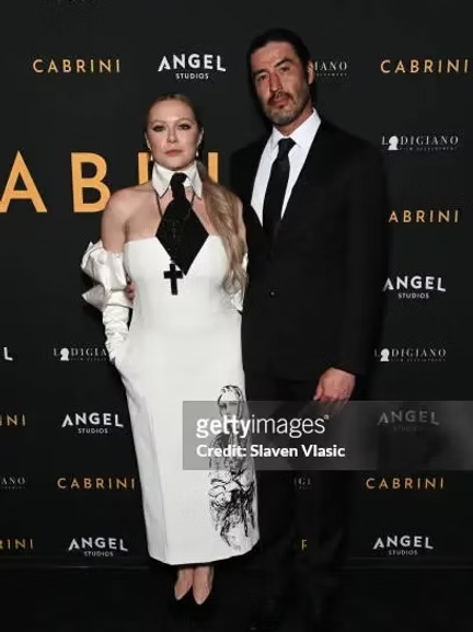 Two people, a woman and a man, dressed formally, standing in front of a black backdrop with gold and white text. The woman is wearing a white dress with black accents and a cross necklace, and the man is wearing a black suit with a white shirt and bl