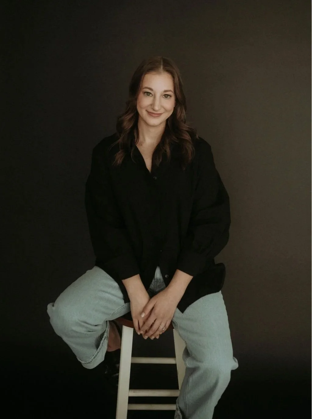 A woman with wavy brown hair sitting on a white stool against a dark background, wearing a black shirt and light gray pants.