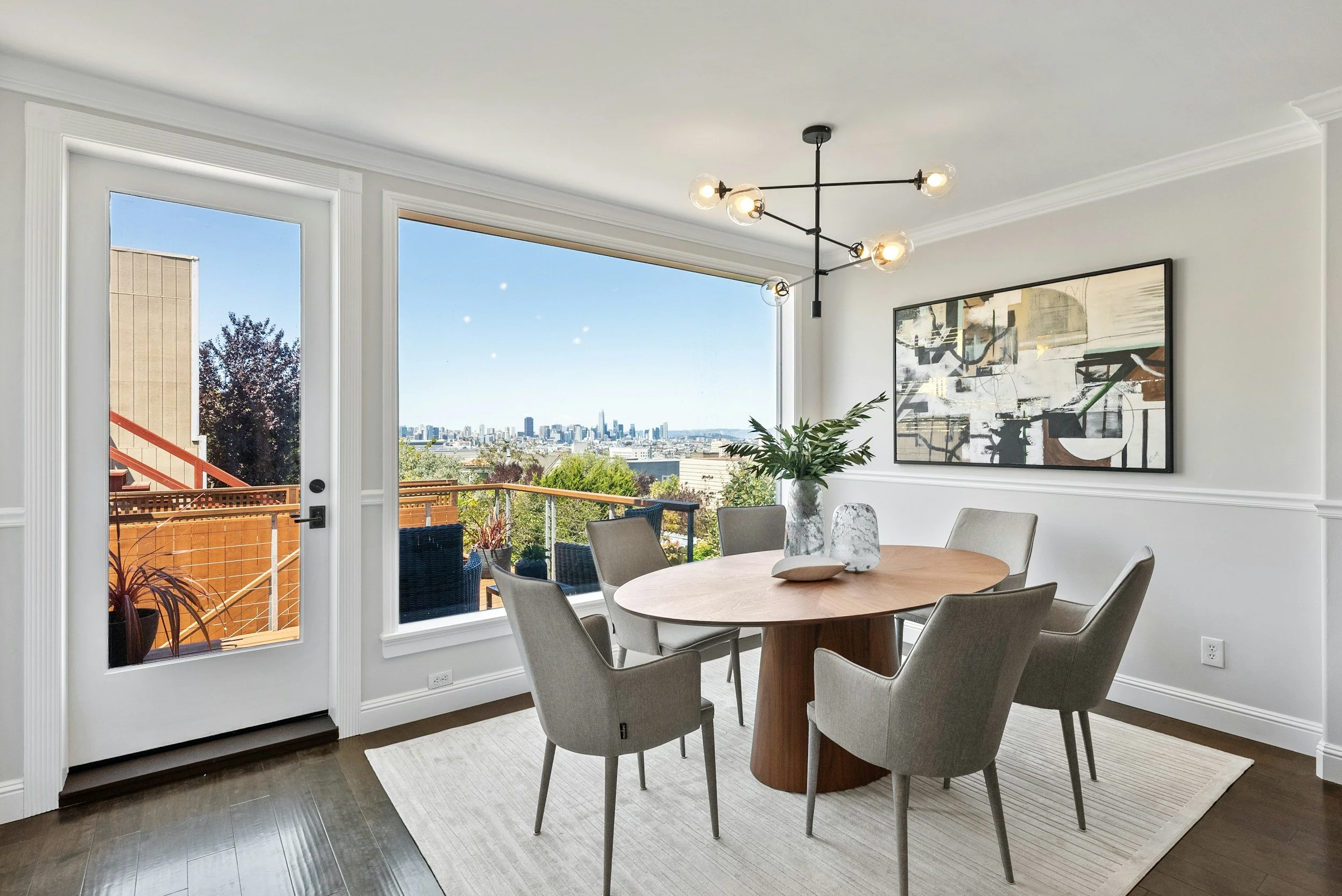 Modern dining room with a round wooden table, six beige chairs, a large window showing cityscape view, a white wall with a large abstract painting, a black chandelier, and a glass door leading to a balcony with outdoor seating.