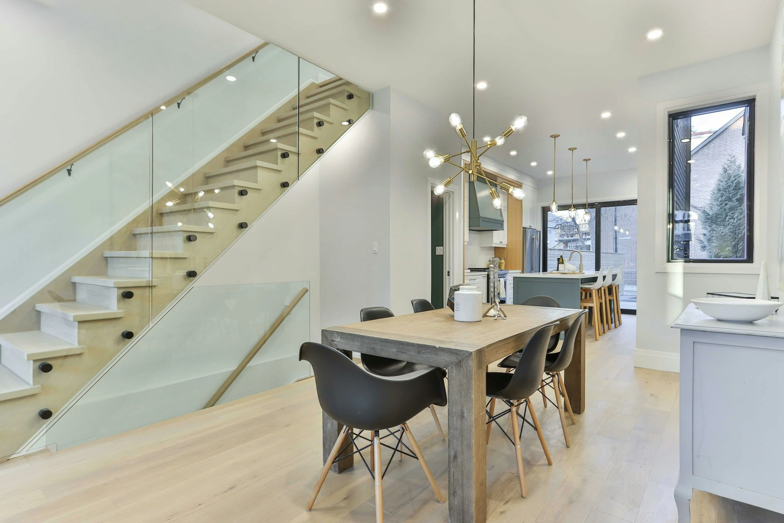 Modern open-concept kitchen and dining area with large windows, a wooden dining table, black chairs, and a kitchen island with bar stools, illuminated by contemporary pendant lights and a chandelier.
