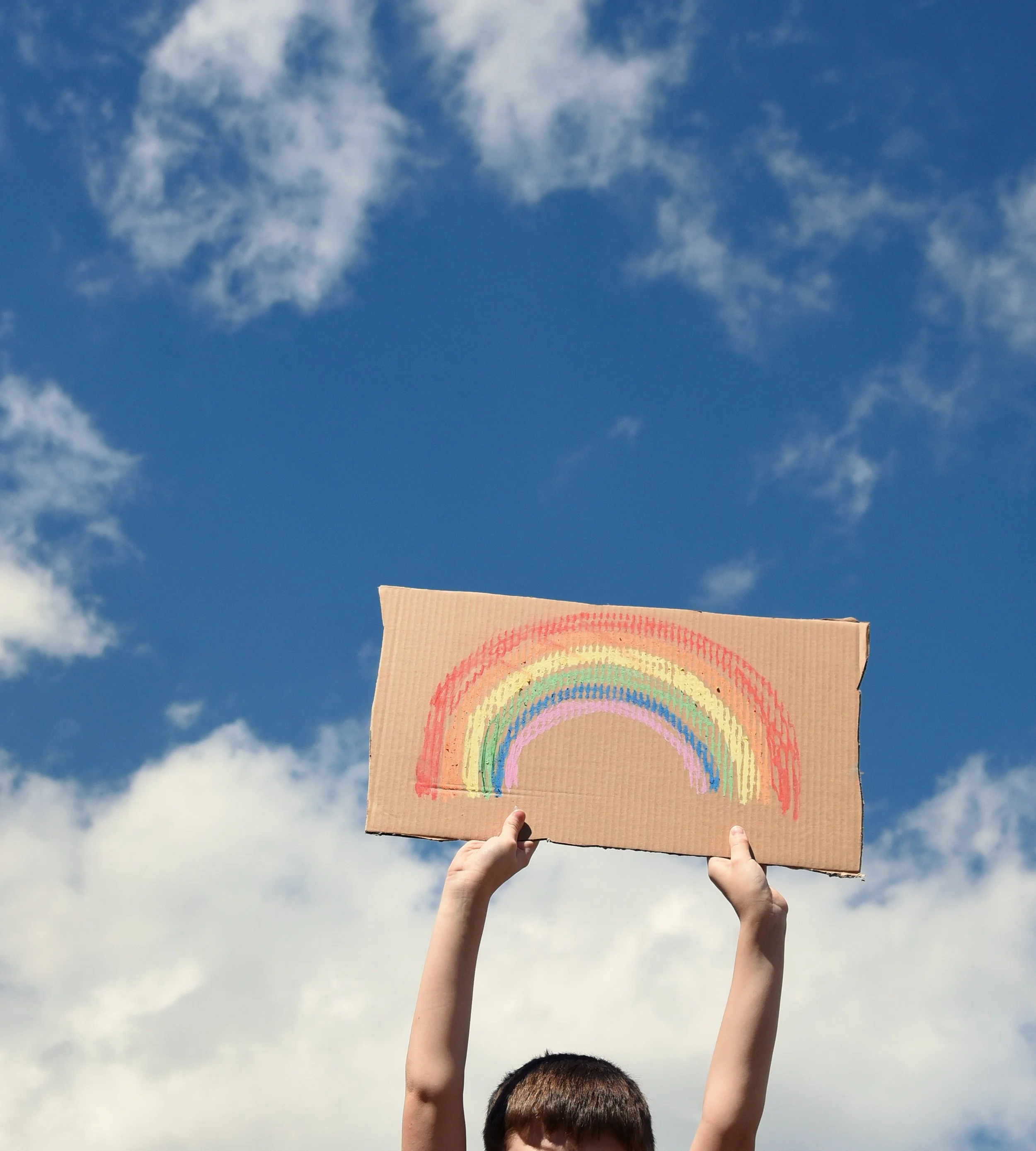 Person holding a cardboard sign with a rainbow drawing against a partly cloudy sky.