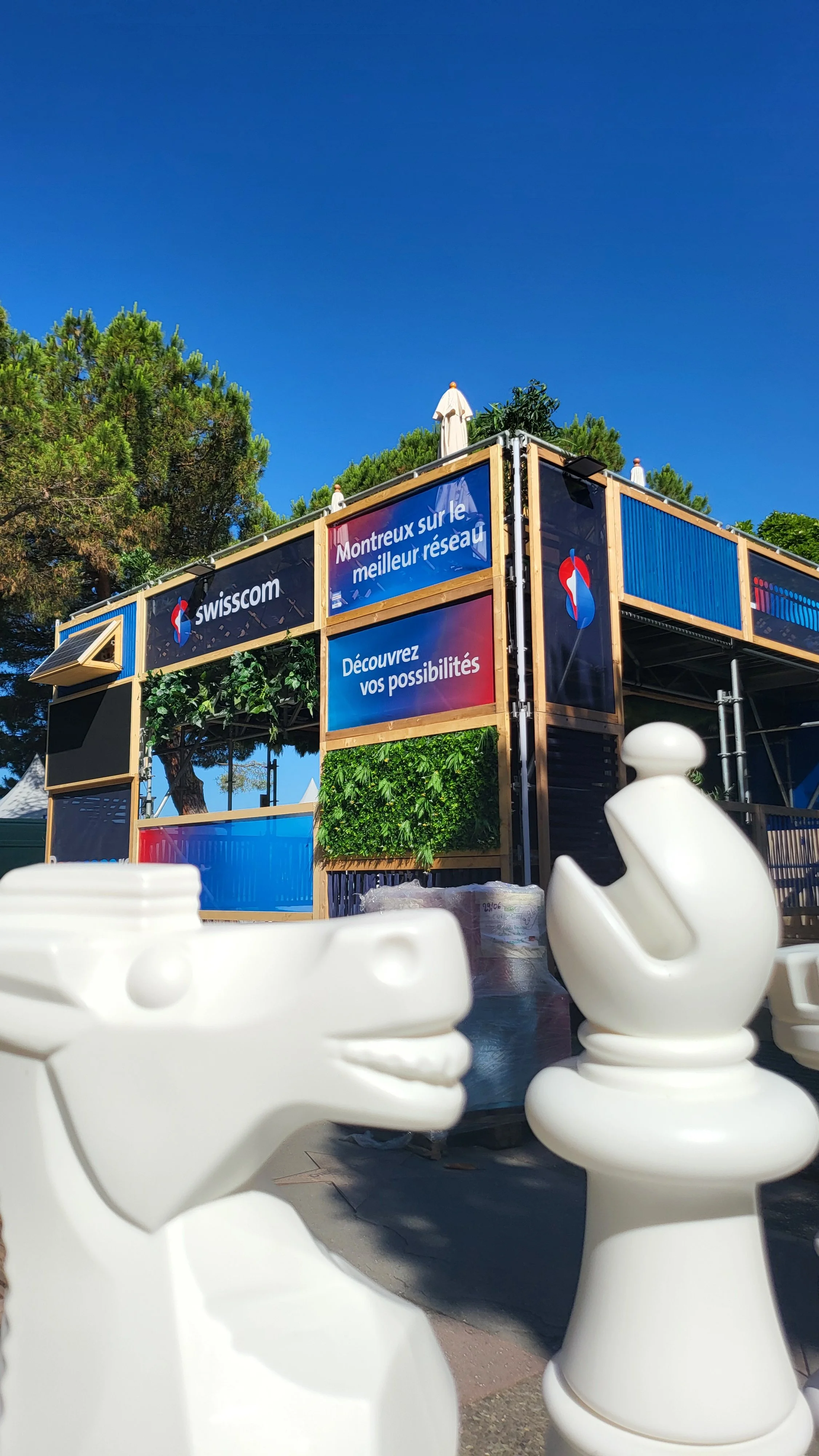 White chess set pieces on a table in front of a promotional stand with Swisscom branding and French text, outdoor setting with trees and a clear blue sky.
