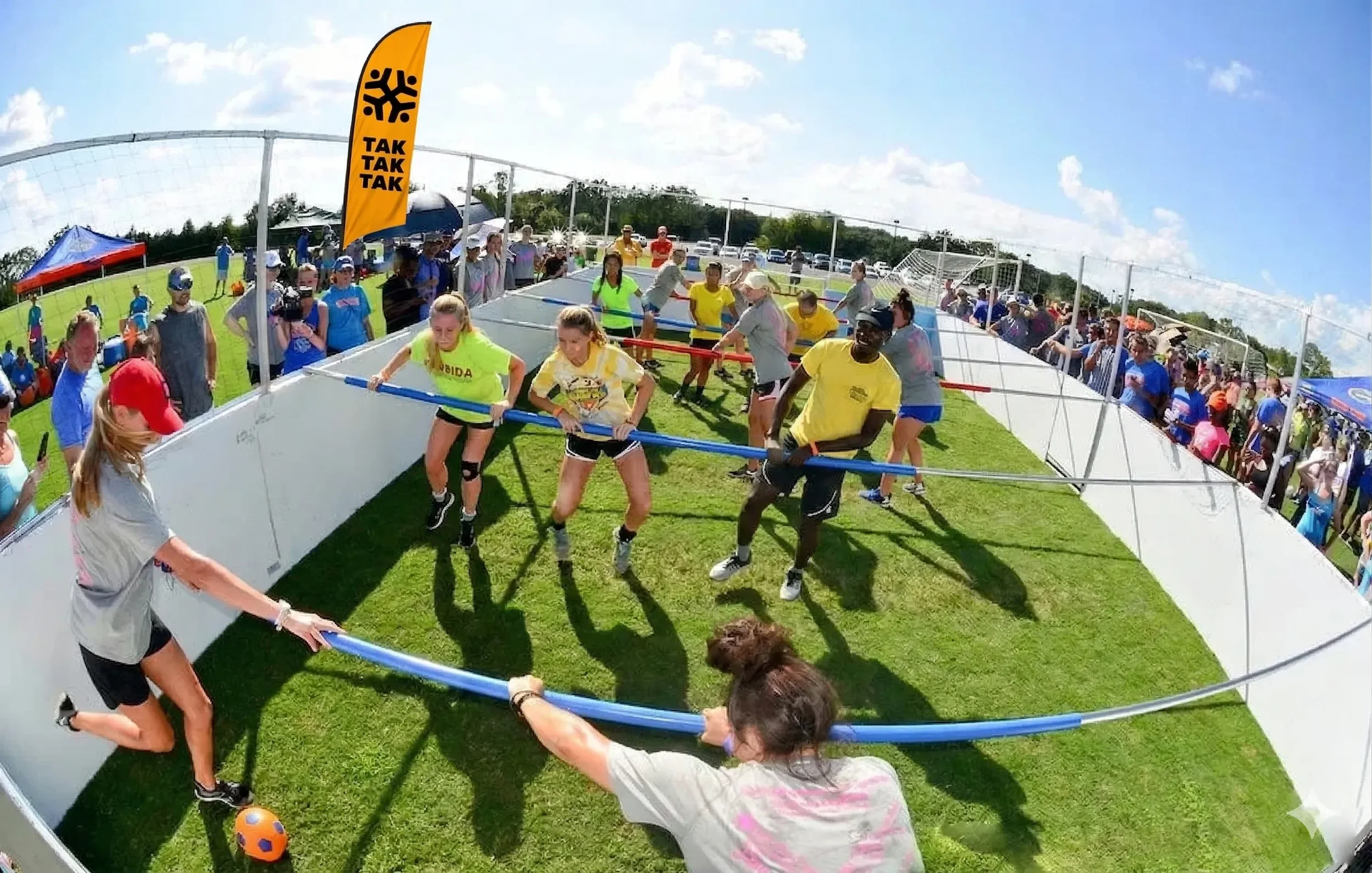 People playing a game of human foosball on a grassy field with a crowd watching behind a white barrier, under a partly cloudy sky.