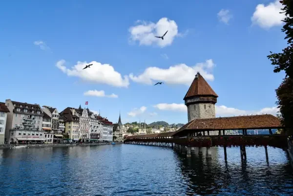 View of a river with historic buildings and a covered wooden bridge in Lucerne, with seagulls flying in a partly cloudy sky.