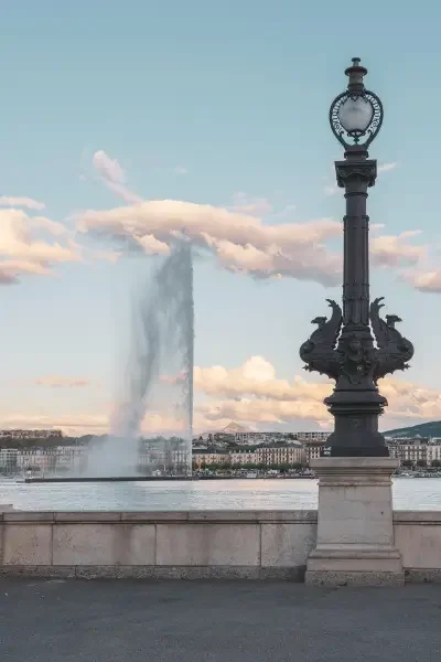 A decorative street lamp next to a waterfront with a large fountain and cityscape in the background.
