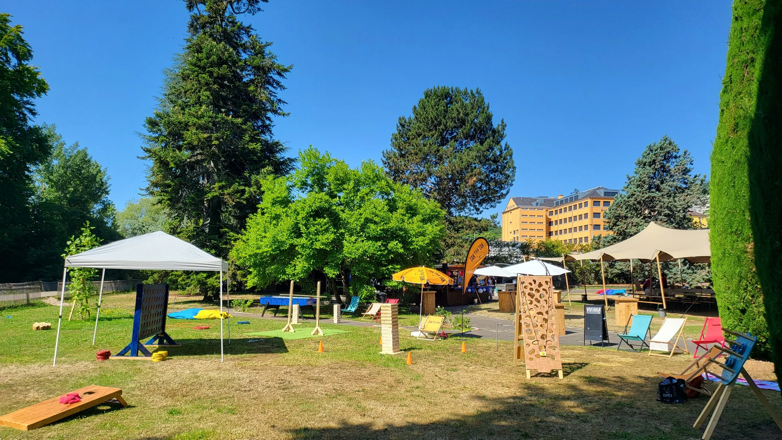 An outdoor park scene with trees, tents, chairs, and game equipment on a sunny day.