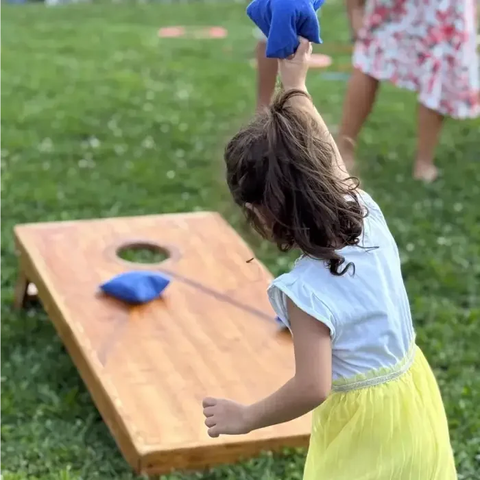 Young girl wearing a yellow skirt and white top playing cornhole outdoors on a grassy field.