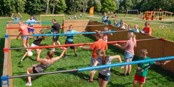 Children playing a game of human foosball outdoors on a grassy field, with some children holding onto horizontal bars and others kicking a soccer ball.