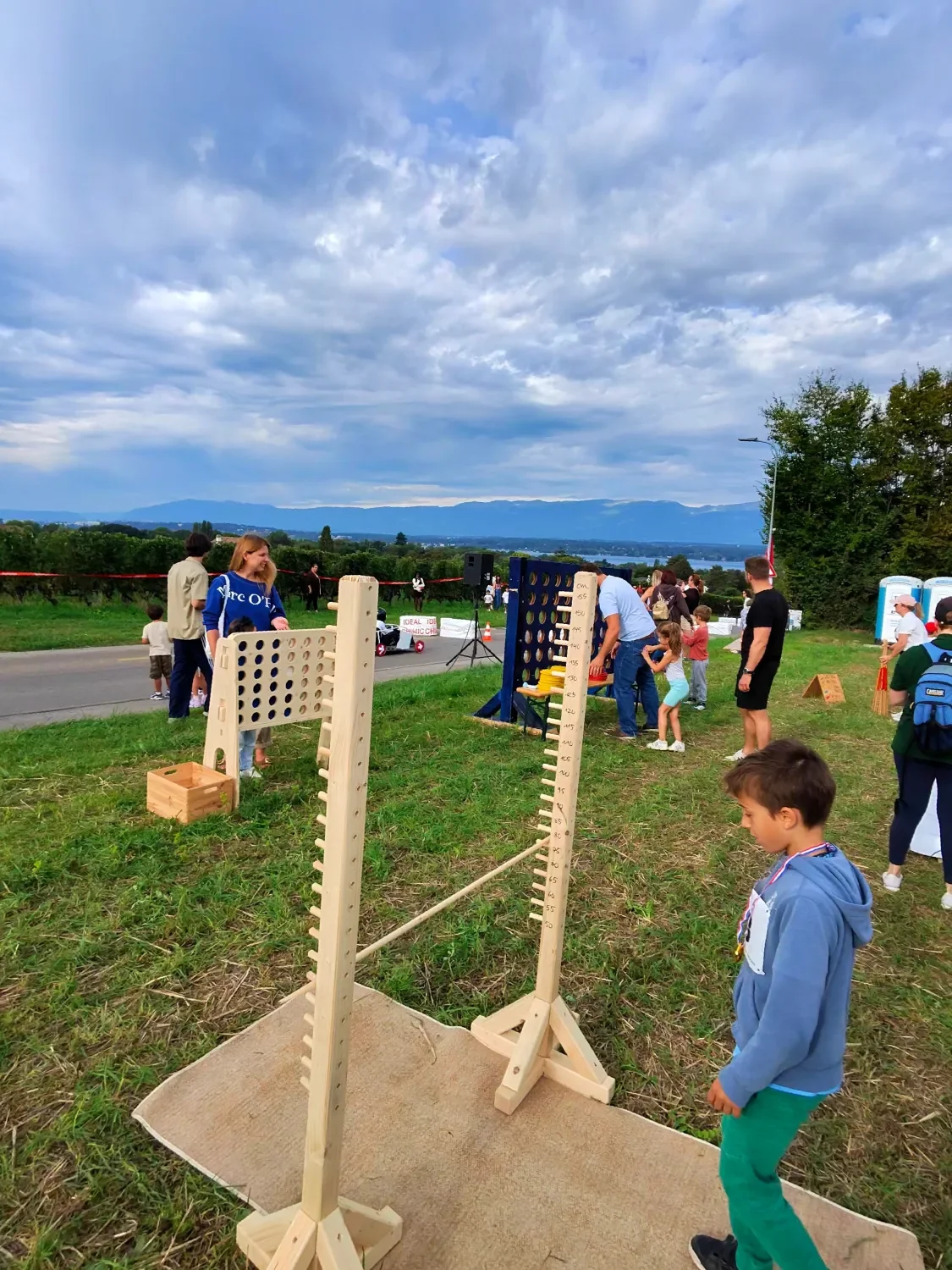 Children and adults playing giant Connect Four and other outdoor games at a park event, with a scenic view of mountains and a body of water in the background.
