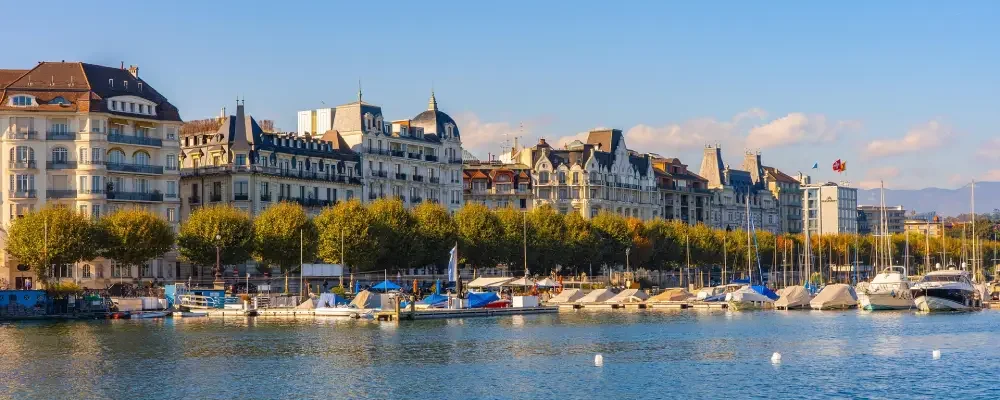 Cityscape of historic Geneva buildings along a waterfront with boats and a row of trees in the foreground.