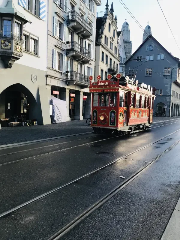 A vintage red tram in Zurich, traveling along wet tram tracks in an urban area with European-style buildings.